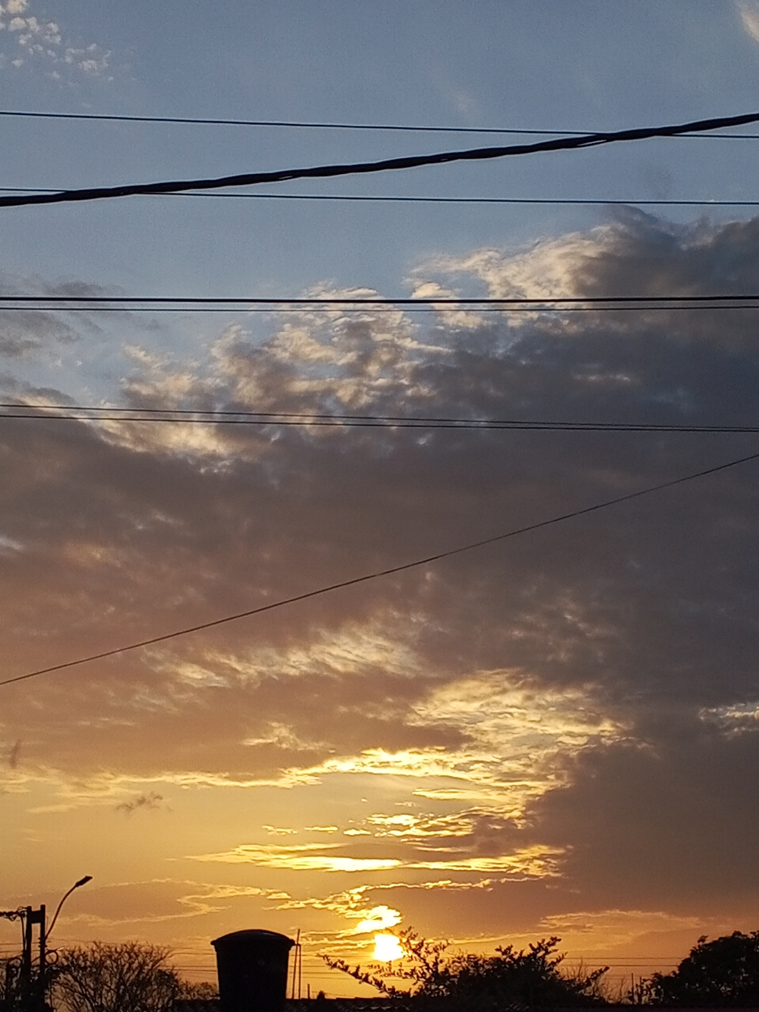 The image depicts a sunset in the city of Unknown, Unknown. The sky is filled with a mix of dark and light clouds, with the sun shining brightly through a gap near the horizon. Power lines are visible in the upper portion of the frame. Silhouetted against the sunset are trees, a street lamp, and a water tank.