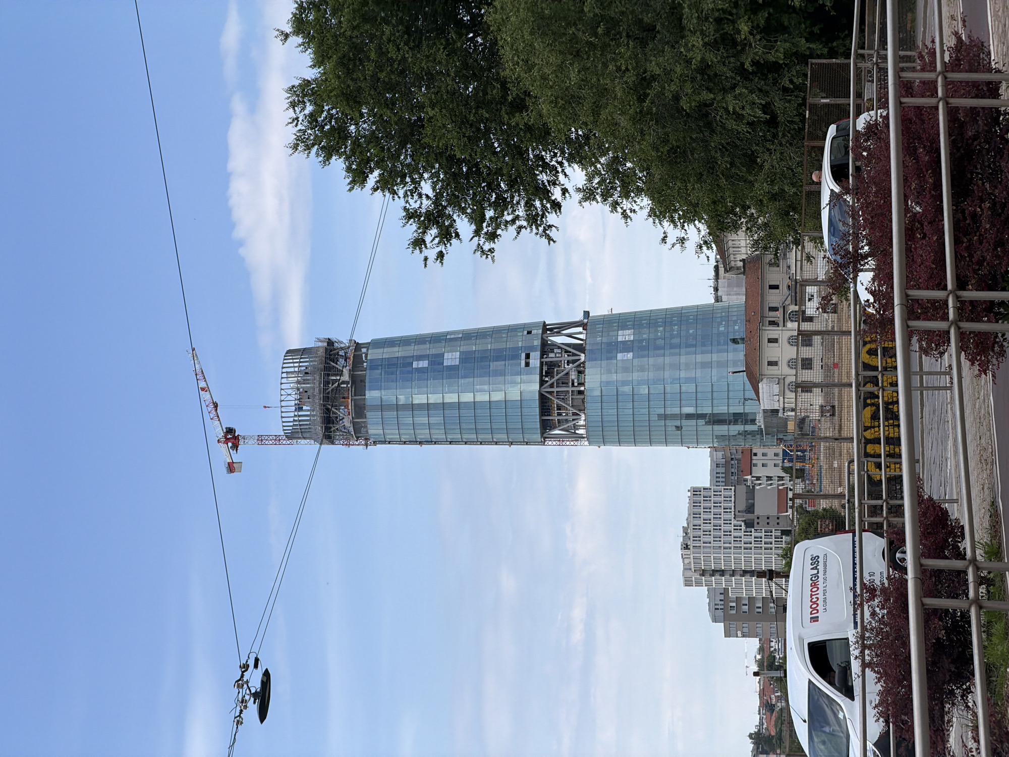 The image shows the Allianz Tower in Milan, Italy, under construction. The tower, a glass skyscraper, is the main subject, with a construction crane visible on its left side. The sky is blue with some clouds. In the foreground, there's a "DOCTORGLASS" van parked next to a fence with bushes. Other buildings can be seen in the midground, giving context to the urban environment of Milan. The scene appears to be taken from a street-level perspective.