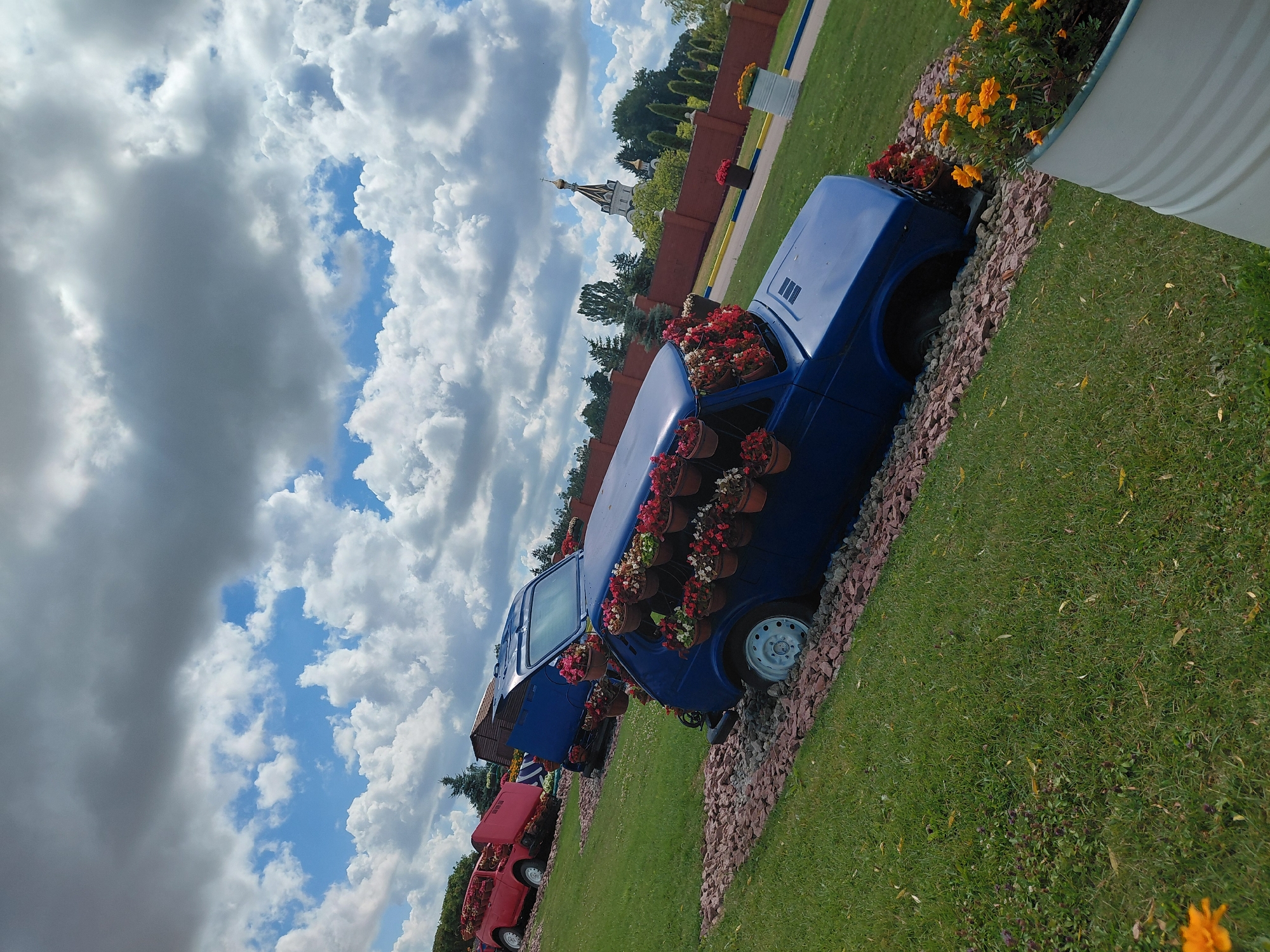 In Kovalivka, Ukraine, an outdoor scene features two vintage vehicles repurposed as planters. A blue car is positioned at a downward angle, with its interior and exterior adorned with numerous potted flowers. In the background, a red car, also filled with flowers, is visible on a slight incline. The cars are situated on a grassy area interspersed with red gravel. Beyond the cars, a brick wall encloses a garden area with trees and a distant church with a golden dome. The sky is filled with dramatic clouds.