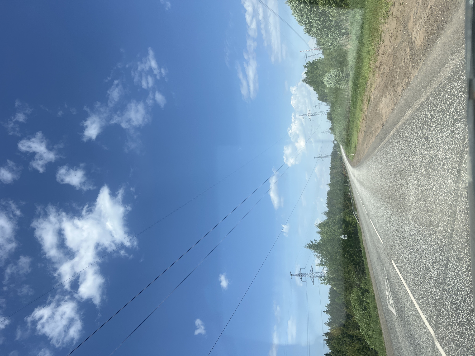 The image depicts a road stretching into the distance under a blue sky with scattered white clouds. Power lines run parallel to the road. On either side of the road are trees. The photo appears to be taken from inside a vehicle, possibly a car. The location cannot be determined as Chaikovsky, Russia without additional information.