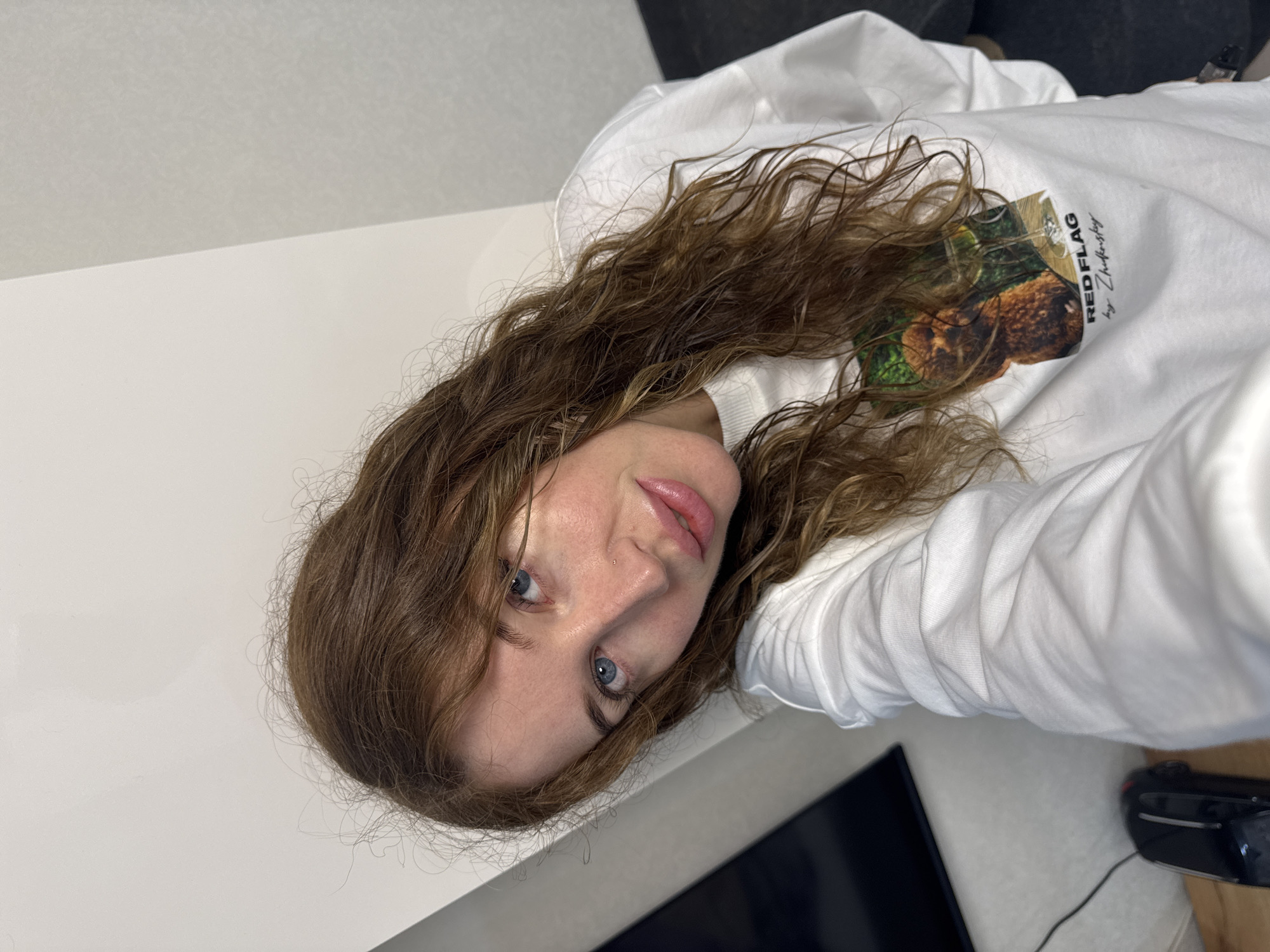 The image depicts a female individual situated in an indoor environment within the city of Chaikovsky, Russia. The subject has long, wavy, reddish-brown hair and blue eyes, which are directed at the viewer. Her complexion is fair, and she has a small, light-colored stud piercing on her right nostril. Her lips are full and appear glossy. She is wearing a white long-sleeved sweatshirt. The front of the sweatshirt features a square graphic depicting a brown, fluffy dog seated at a table alongside a wine glass containing a pale liquid. Below this graphic, the text "RED FLAG by Zhukovsky" is printed in black font. The background consists of light-colored walls, with a vertical seam or corner visible, showing a slight tonal difference between two wall sections. To the left, a dark rectangular object, possibly a screen, is partially visible, along with a black electronic device resting on a wooden surface connected by a cable. The subject's pose suggests a self-portrait or direct address to the camera.
