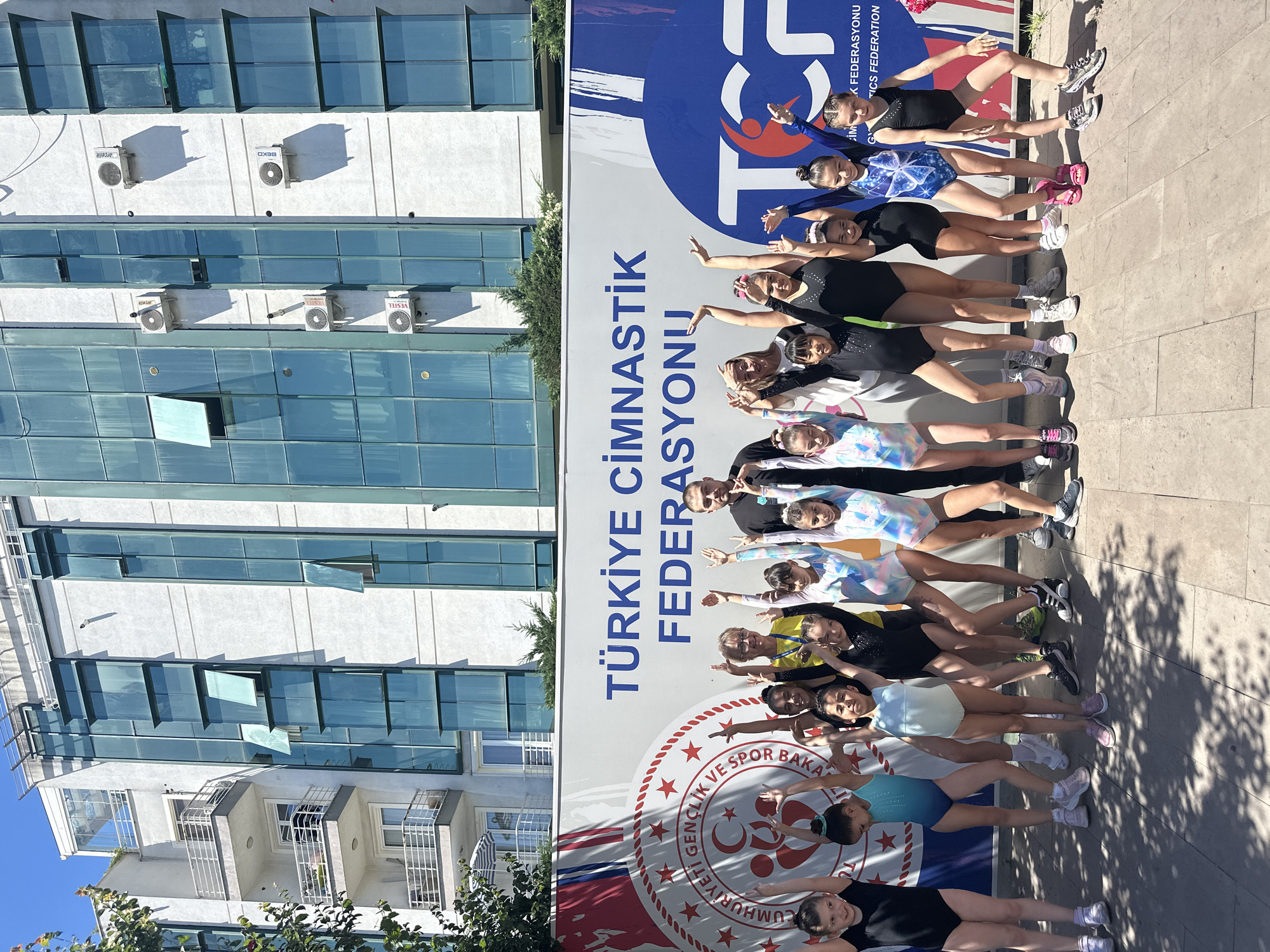 In the city of Sancaktepe, Turkey, a group of young female gymnasts, aged approximately 6 to 12 years old, pose for a photograph with two adults in front of a large banner. The banner features the text "TÜRKİYE CİMNASTİK FEDERASYONU" (Turkish Gymnastics Federation) and the logo of the federation.
The gymnasts are dressed in various leotards, some plain black, others patterned in bright colors. They are standing in two rows, with their arms raised in the air. The adults, a man and a woman, are standing behind the children. The woman is wearing a yellow top and glasses, while the man is wearing a black shirt.
The background consists of a modern white building with large, tinted blue windows and several air conditioning units mounted on the exterior wall. The building appears to be multi-story with balconies visible on the upper floors. The ground in front of the banner is paved with light-colored stones, and shadows from overhead foliage are cast on the ground. The overall scene suggests an outdoor event related to gymnastics.