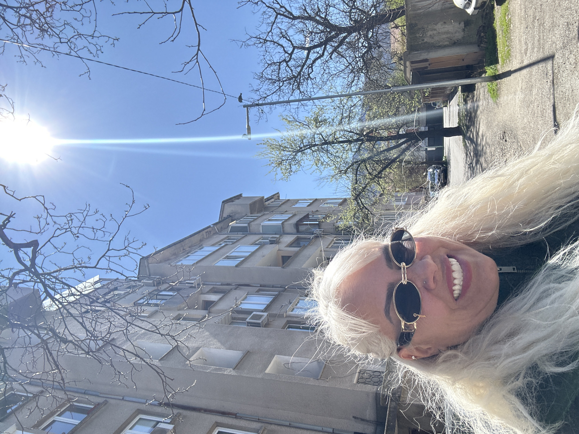 A low-angle, outdoor shot shows a woman with blonde hair and sunglasses. Her head is tilted to the right, and she is smiling, revealing her teeth. Behind her, a multi-story building with many windows and air conditioning units is visible. A street light with a bird perched on top is in the background. Bare tree branches are partially obscuring the view of the building and the blue sky with a bright sun. A narrow street is to the right.
There is no definitive information to identify the specific location as Sofia, Bulgaria. Additional visual cues or metadata would be necessary to confirm.