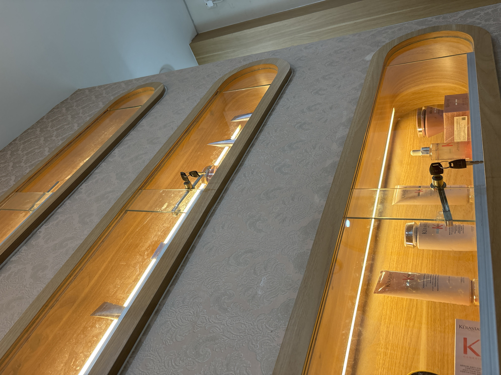 Here is a forensic description of the image:
Three vertical, arched display cases are recessed into a light gray wall with a subtle damask pattern. The cases are constructed of light-colored wood and contain glass shelves, with products displayed inside.
The first display case on the left contains various objects, including what appears to be a set of keys attached to an item. The display case in the middle holds similar items, possibly also sets of keys.
The display case on the right features a variety of Kerastase hair care products. These are arranged on the glass shelves, with tubes, bottles, and product packaging visible.
The location is likely a salon in North Jakarta, Indonesia, given the types of products displayed and the background setting.