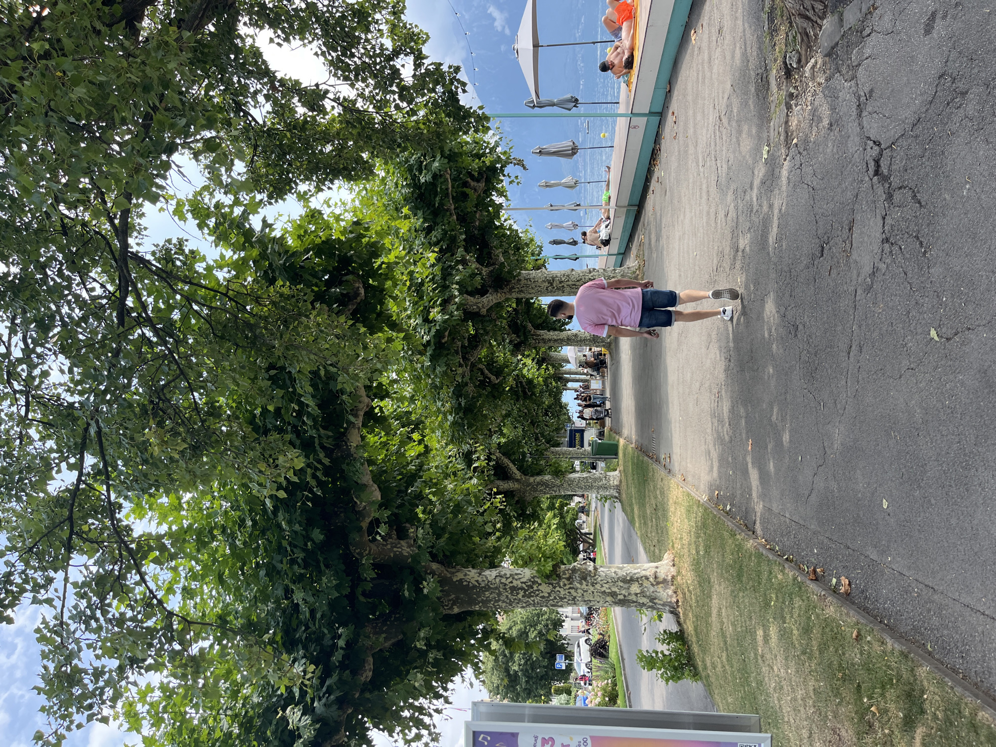 The image depicts a scene along the waterfront promenade of La Tour-de-Peilz, Switzerland. A man wearing a pink t-shirt and blue shorts walks away from the camera on a paved pathway. The pathway is bordered by a grassy area on the left and a body of water on the right. Large trees with textured bark line the pathway, providing shade. On the right side of the pathway, people are relaxing on orange towels near the water's edge, some lying down and others sunbathing. Beach umbrellas are set up along the water. In the background, a mountain range can be seen across the water, shrouded in light clouds. The sky is partly cloudy with patches of blue. A street with parked cars and pedestrians is visible to the left of the trees. A lamppost with some signage is also present on the far left.