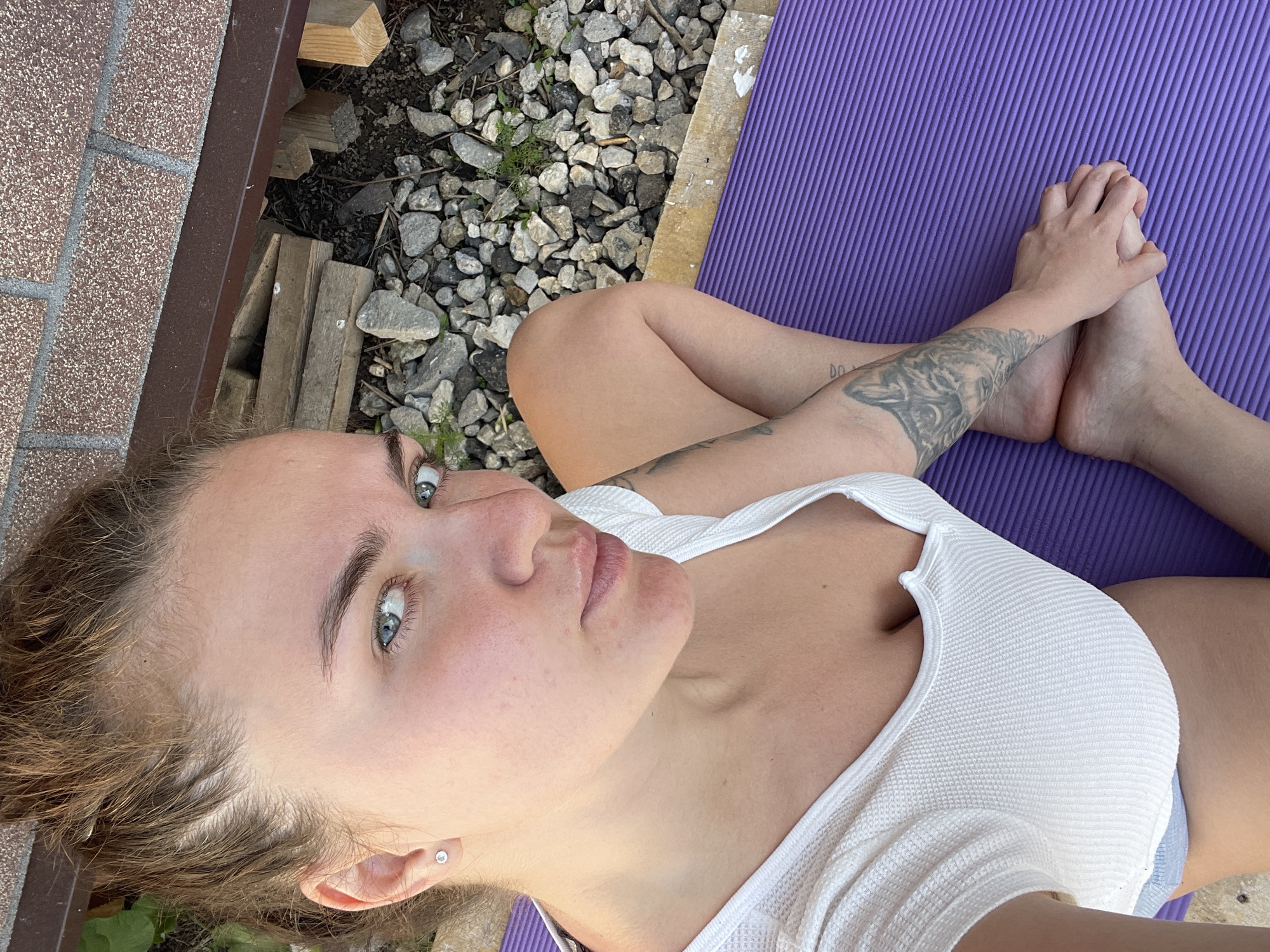 A woman is lying on a purple yoga mat outdoors. Her legs are crossed, and her hands are clasped around her feet. She is wearing a white top. There are pebbles and some greenery beside the mat, and a wooden structure.
The location is Мосметаллоконструкция Онт, Russia is not discernible.