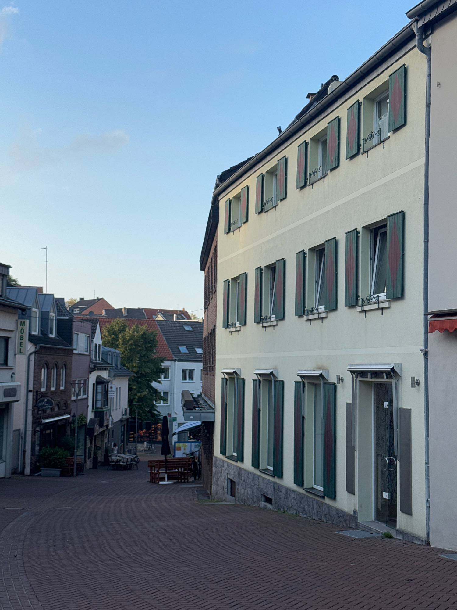 A narrow, cobblestone street winds through a European city, lined with multi-story buildings. The buildings feature traditional architecture with pitched roofs and windows adorned with green shutters. One building on the right side of the street is painted a light yellow and has a row of open windows with red diamond patterns on the green shutters. A glass door with a metal handle is visible on the ground floor of this building. On the left side of the street, other buildings display a variety of colors and architectural styles, including some with signs. The street is mostly empty, with some outdoor seating areas visible, including tables and benches with an umbrella. The sky is clear blue with a few wispy clouds. The scene appears to be set in the late afternoon or early evening. Based on the architectural style and the sign "MÖBEL" (furniture in German) visible on a building on the left, this location is likely in Cleves, Germany.