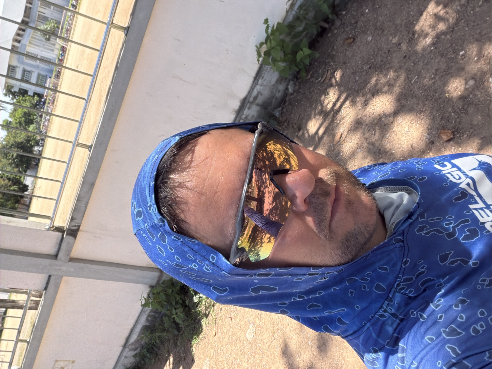A man wearing a blue hooded shirt with a light blue pattern and sunglasses is taking a selfie. He has short brown hair and stubble. The sunglasses reflect trees and a building. The background shows a white wall, a metal fence, a dirt path, and vegetation. In the distance, there is a building complex and a sports field. The image appears to have been taken outdoors during the day. The location is the city of Baan Bo Phut, Thailand.