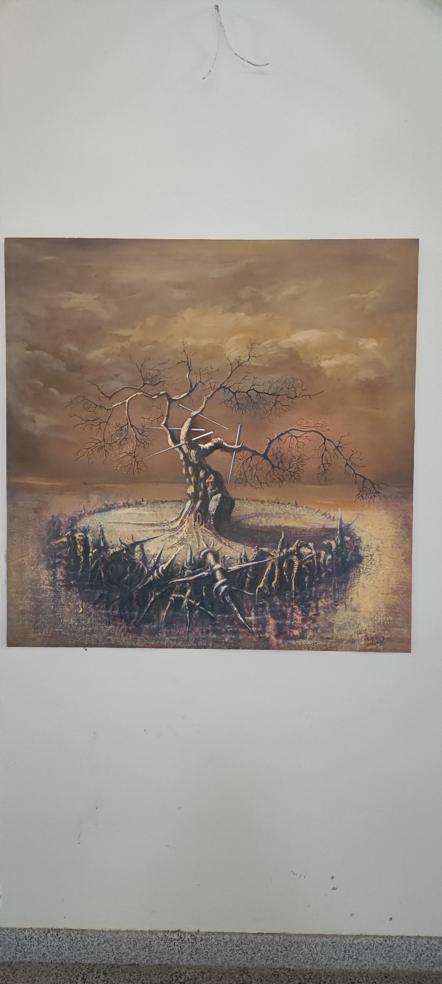 The image is a painting depicting a gnarled, leafless tree at its center. The tree's trunk appears to be made of stone or rock, and it has several sharp, metallic objects embedded in its branches and trunk. Surrounding the base of the tree is a circular formation of numerous figures, which appear to be human or humanoid, with pointed, spike-like protrusions from their heads and bodies, creating a defensive or menacing perimeter. The ground around the tree and figures is textured and appears somewhat desolate. The sky is overcast with swirling clouds in shades of brown and orange, suggesting a dramatic or somber atmosphere. The painting is framed by a white wall and the floor is visible at the bottom of the image. This artwork is located in the Voskevaz Children's Art School in Voskevaz, Armenia.