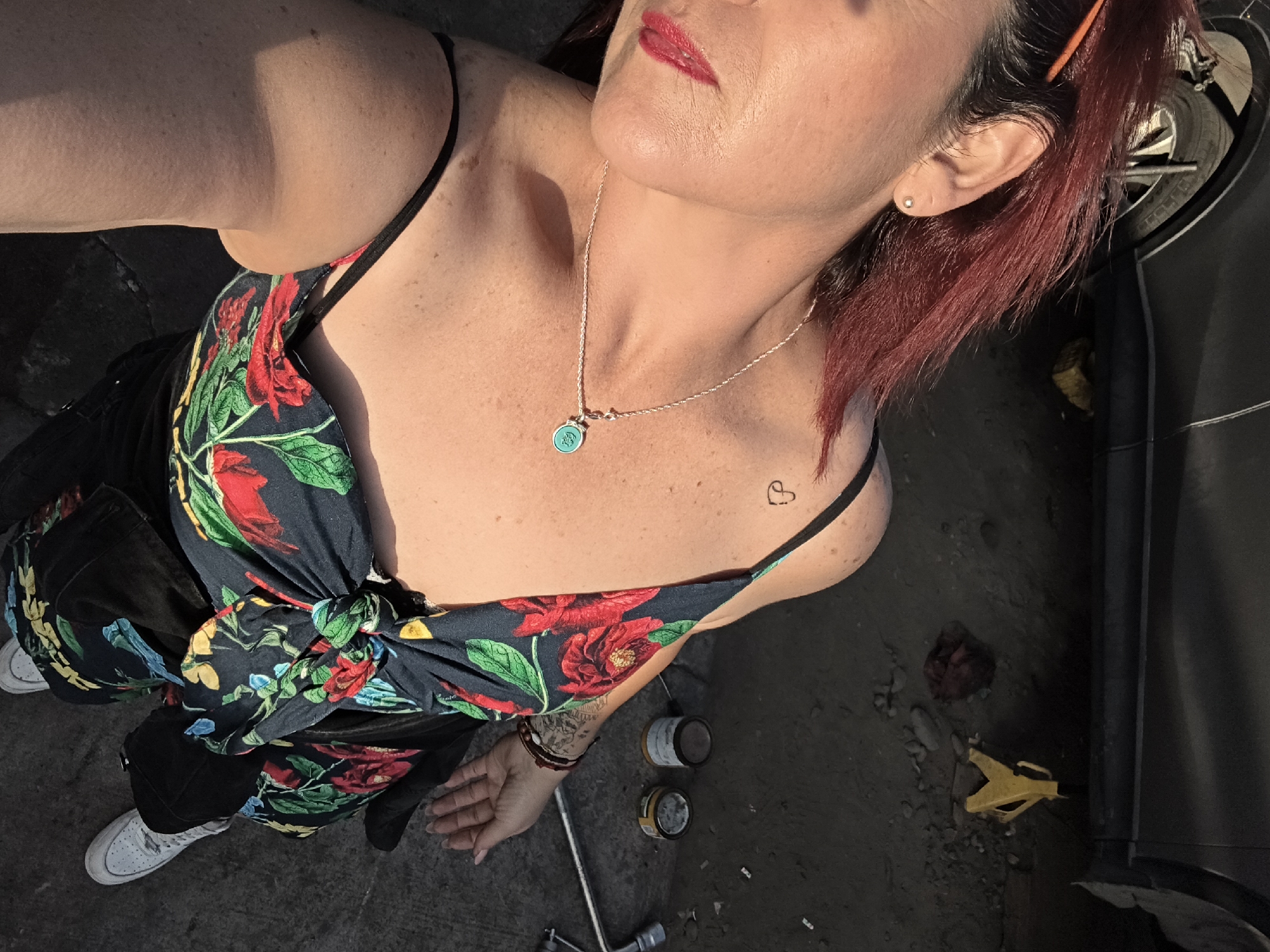 A woman with red hair and wearing a floral-print top, black pants, and white sneakers is seen from an overhead angle. She has a necklace and a small heart tattoo on her upper left chest. The location appears to be a garage or workshop, with a car, tools (including a lug wrench and a jack), and paint cans visible on the ground. The light suggests it is outdoors or near an open entrance. The specific location within Arica, Chile cannot be determined from the image.