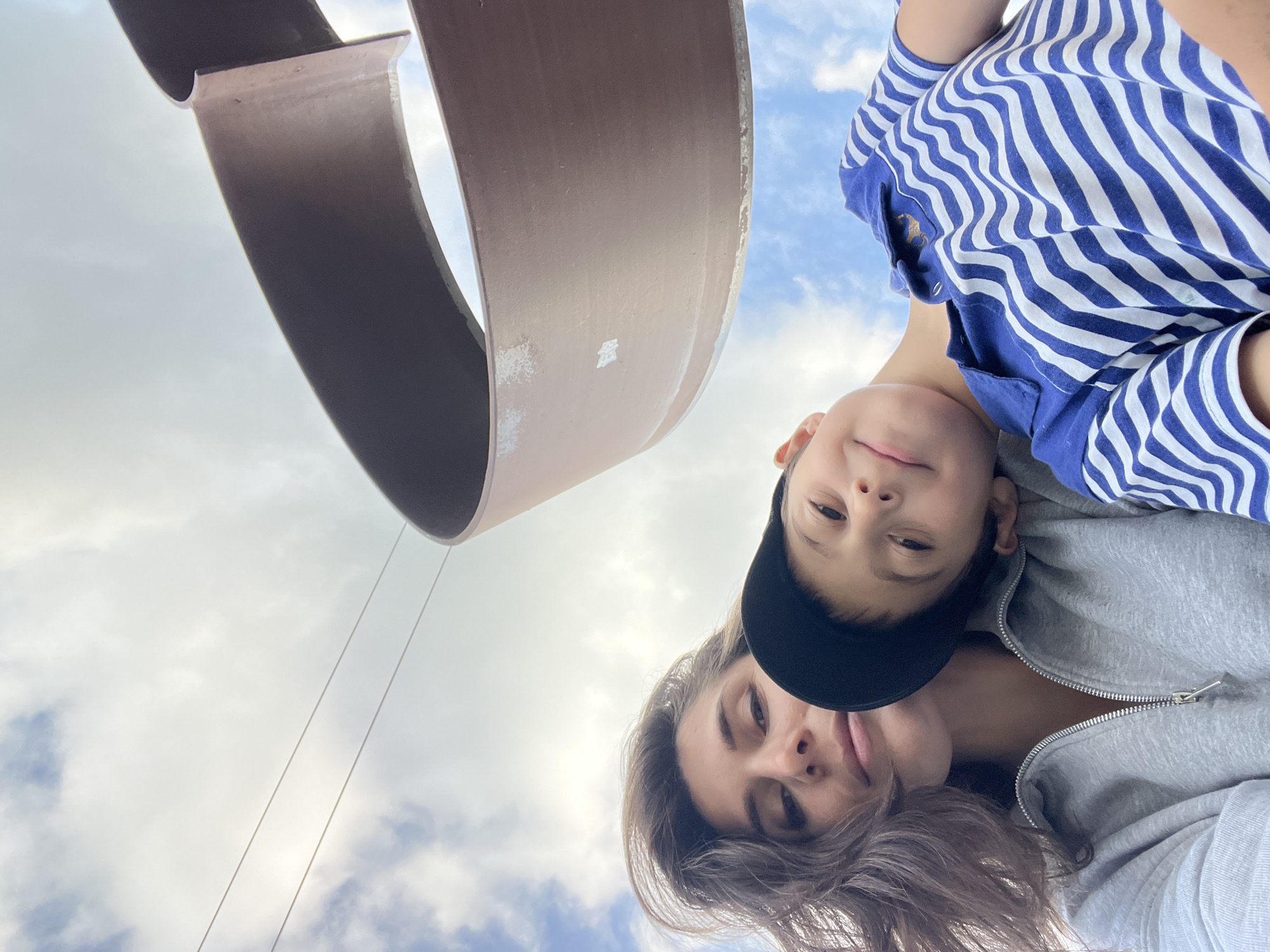 In a low-angle selfie, a woman and a young boy are pictured against a cloudy sky, with a large, abstract metal sculpture dominating the upper right portion of the frame. The woman, with dark, wavy hair and a neutral expression, wears a grey hooded sweatshirt. The boy, with a black baseball cap on backward and a blue and white striped polo shirt, rests his head on the woman's shoulder, smiling towards the camera. Thin wires, possibly for a cable car or power lines, are visible against the sky. The sculpture appears to be made of dark, reddish-brown metal with a curved, ribbon-like form. The setting is outdoors, and the sky suggests it may be daytime. The presence of a large, unique sculpture and overhead wires could indicate a public park, plaza, or area with a specific architectural feature within the city of Chaikovsky, Russia.