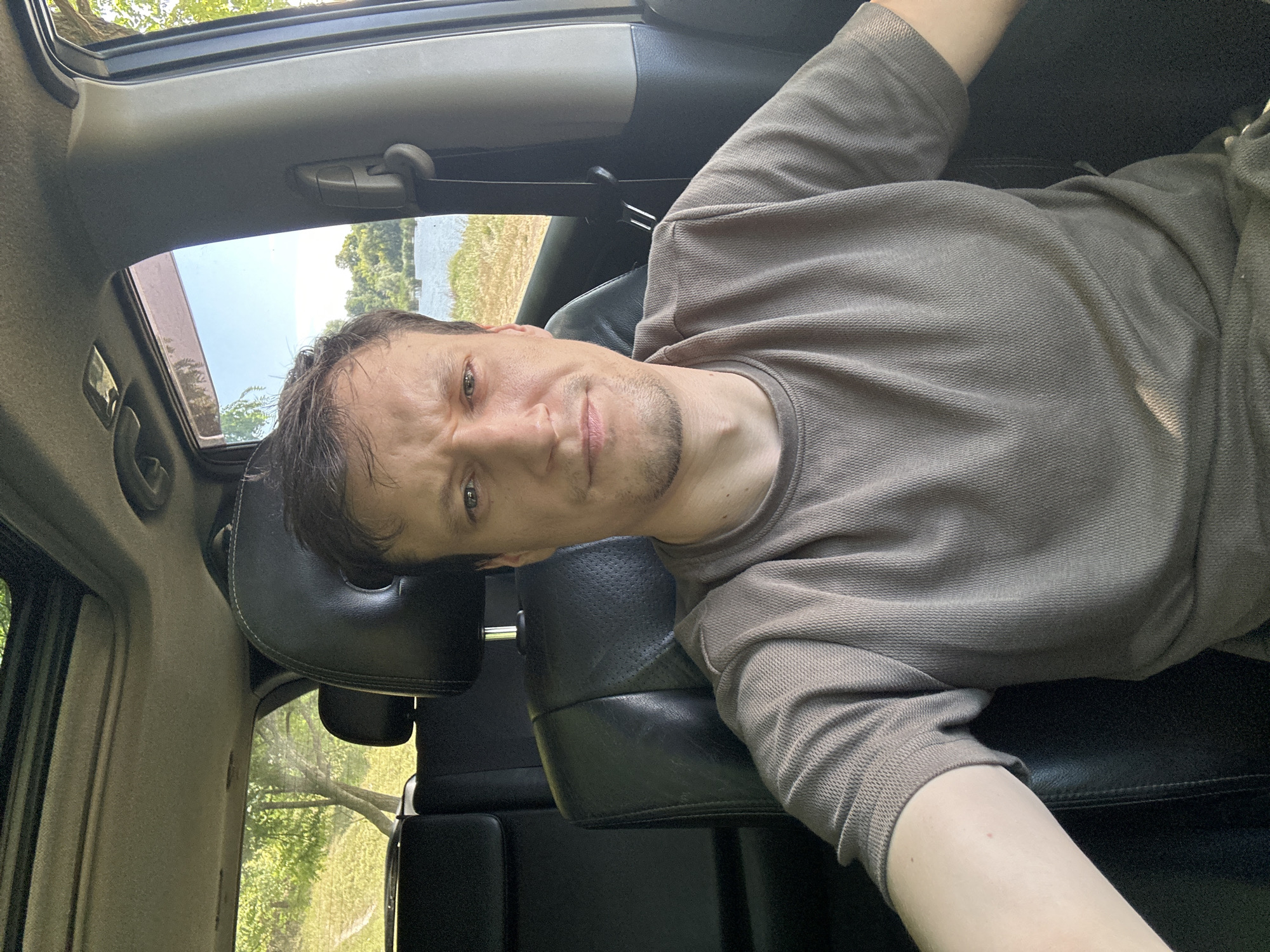 A man with short brown hair and a skeptical expression is sitting in the driver's seat of a car. He is wearing a grey t-shirt with a waffle knit texture. The interior of the car is visible, including black leather seats and a sunroof. Through the windows, a body of water, grassy hills, and trees can be seen, suggesting an outdoor location. The man appears to be looking directly at the camera, possibly taking a selfie. The scene is lit by natural daylight. The image has been cropped in some sections to provide closer views of various parts of the scene. There is no specific information provided in the image that indicates the location is in Makiv, Ukraine.