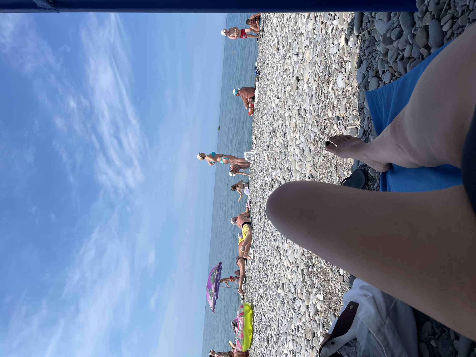 The image depicts a sunny day at a crowded pebble beach. In the foreground, the legs of an individual are visible, resting on a blue towel. Beyond them, the beach is populated with several people sunbathing and relaxing. A purple umbrella provides shade for some individuals. The sea is calm and blue under a sky with scattered white clouds. Several inflatable toys are visible on the beach. The overall scene is one of a typical summer day at the seaside.