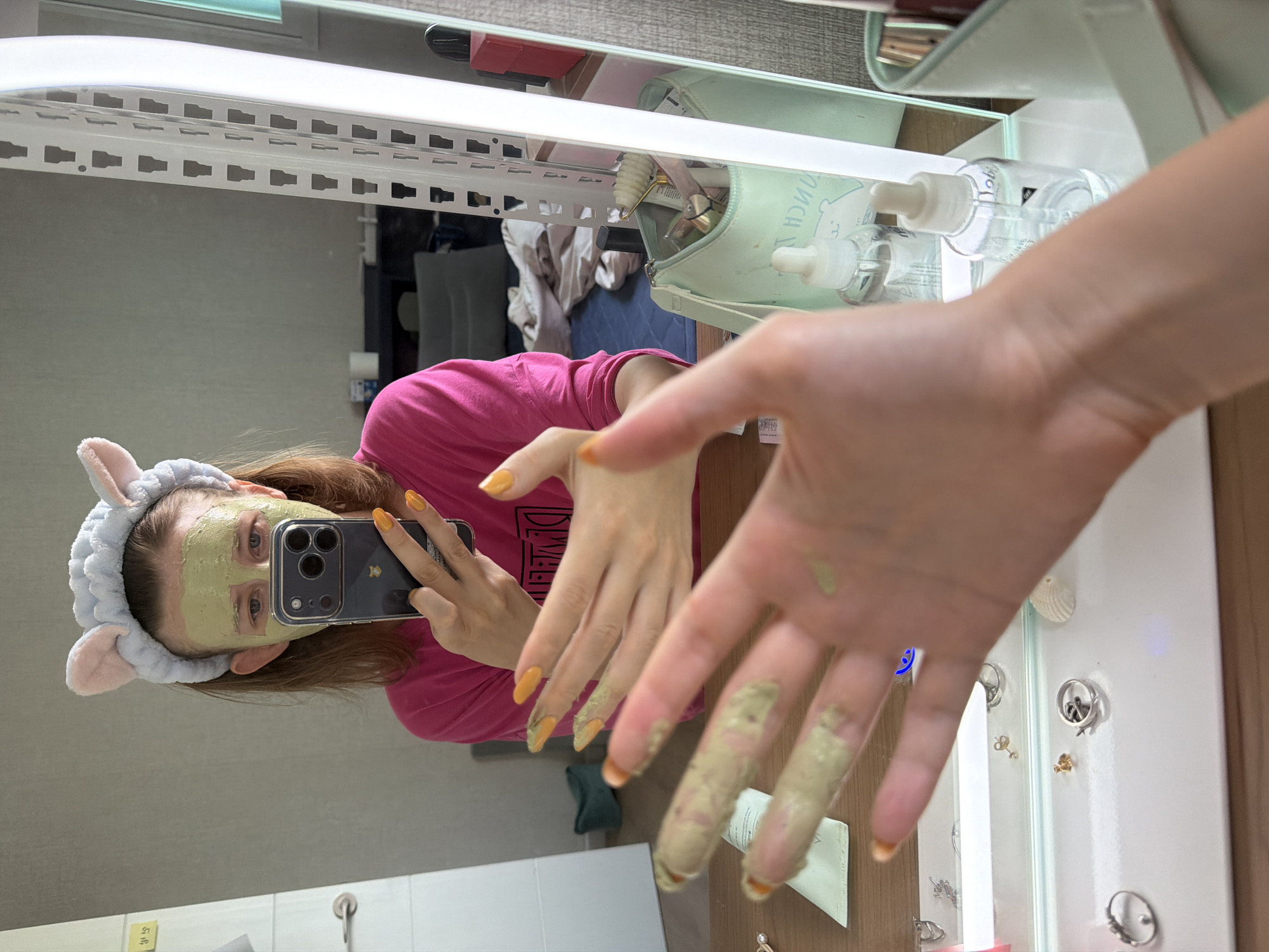 A young woman is taking a selfie in a mirror in a room. She is wearing a pink shirt and a light blue headband with cat ears. Her face is covered in a green face mask. She is holding a black smartphone in her right hand, which is angled to capture her reflection and her face mask. Her fingernails are painted orange. Her left hand is also visible, with green face mask applied to her fingers. In the foreground, her left hand and forearm are in focus, with the face mask clearly visible on her fingers. In the background, reflected in the mirror, is a dresser with various cosmetics and toiletries, including a light green makeup bag filled with items, and a bottle of clear liquid. There is also a bed with rumpled bedding. The overall setting appears to be a personal bedroom. The location is stated to be Dongducheon-si, South Korea.