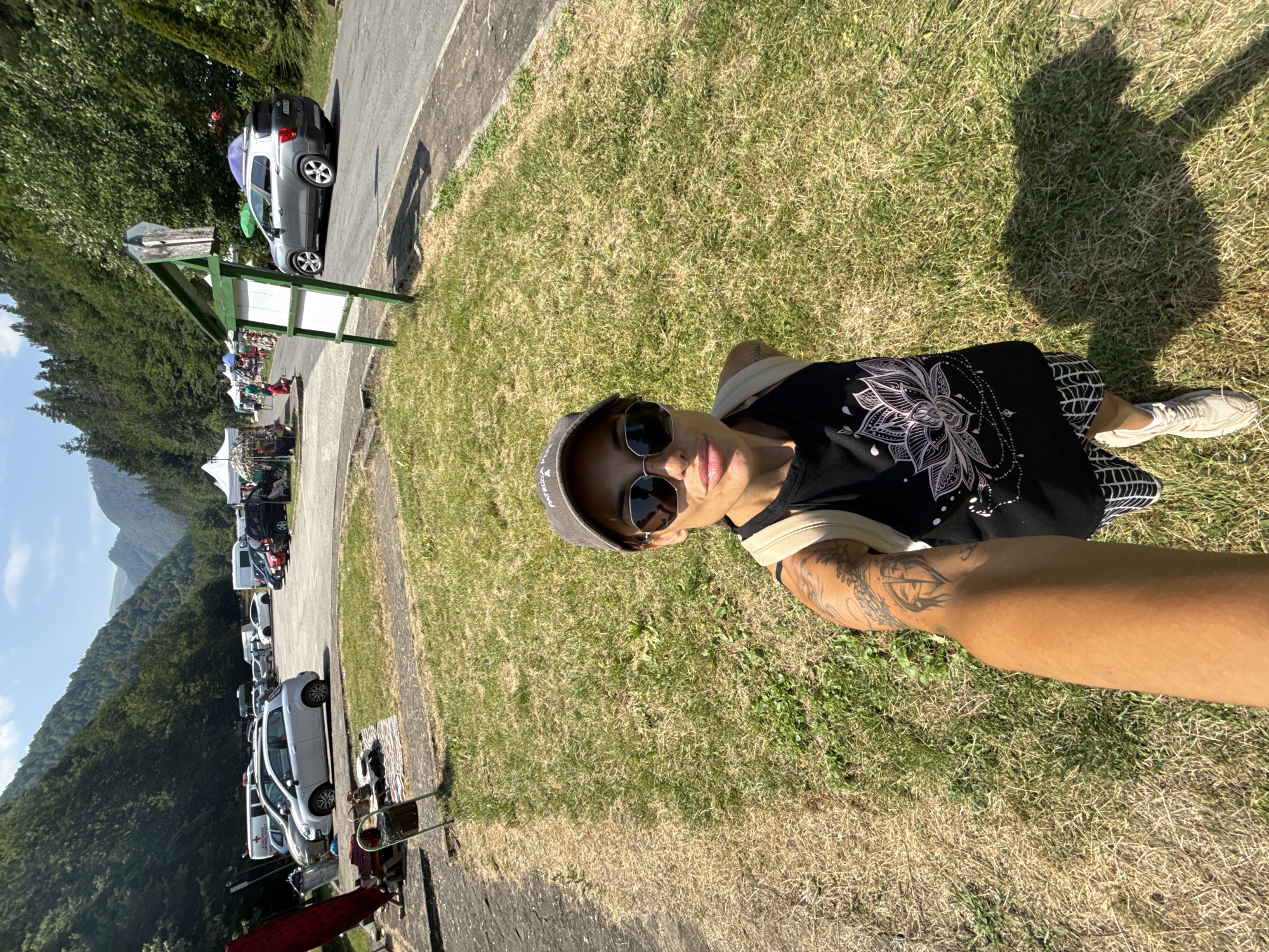 The image captures a selfie taken by a woman in Ribaritsa, Bulgaria, a location characterized by a mountainous landscape with dense green forests. The woman, wearing a black tank top with a lotus flower design, a grey baseball cap, and sunglasses, stands on a grassy area. Her arm, adorned with tattoos, is extended to take the selfie. In the background, a parking lot is visible with several cars, including a silver Fiat Punto and a grey SUV. Tents and stalls suggest a market or a temporary event is taking place. The environment is sunny with a clear blue sky.