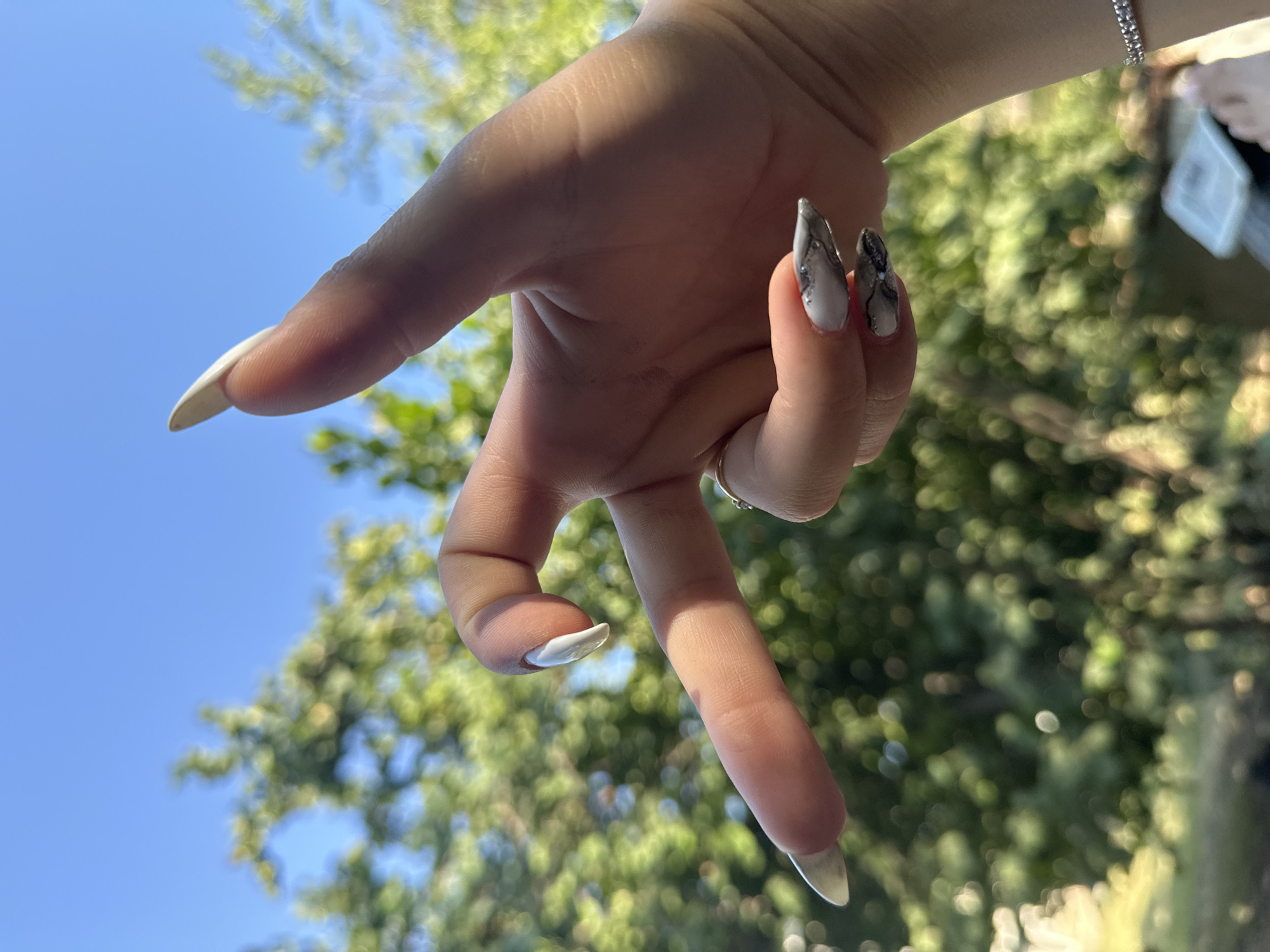 A close-up, outdoor, eye-level shot shows a person's left hand in the foreground, positioned against a blurred background of green foliage and a bright blue sky. The hand is held in a gesture with the index finger extended to the left, the middle finger bent downwards and partially obscuring the ring finger, and the thumb curled upwards. The fingernails are long and pointed, with a white polish on the extended index and middle fingers, and a grey and white marbled design on the nails of the ring and pinky fingers. A thin, silver ring is visible on the ring finger. The image was captured on August 15, 2023, at approximately 1:42 PM.
