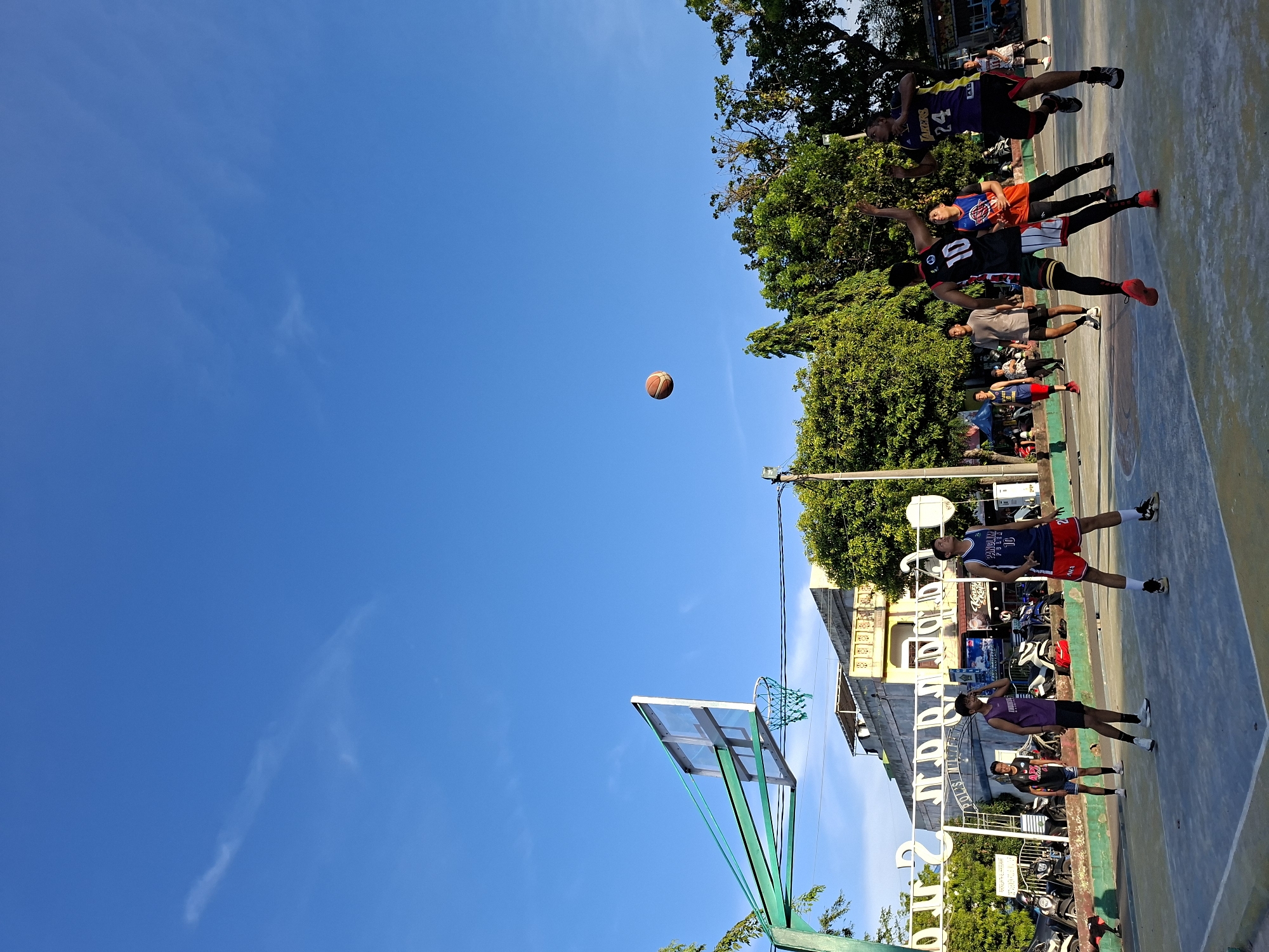 The image depicts a basketball game in Lhokseumawe, Indonesia, under a clear blue sky. A basketball is suspended in mid-air between the players and the hoop. The court is surrounded by trees and buildings. Several players are visible, wearing basketball jerseys numbered 10 and 24. People are standing and sitting along the sidelines. The basketball hoop is green. A building is visible in the background.