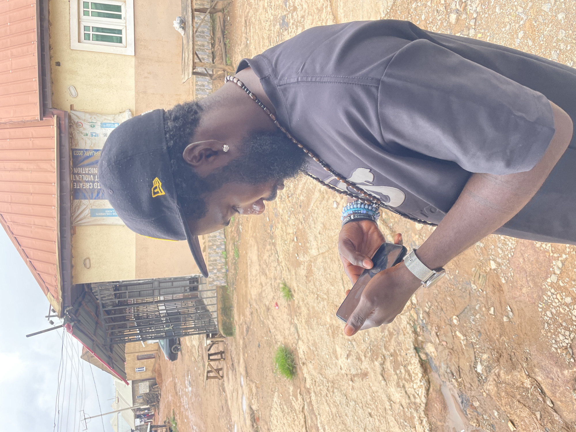 In Kurunduma, Nigeria, a man with a beard and a black baseball cap with a yellow logo stands outdoors, looking down at his phone. He wears a black t-shirt, a silver watch on his left wrist, and multiple beaded bracelets and necklaces. The ground is dirt with patches of grass and puddles, and in the background, a building with a red roof and a metal-barred enclosure is visible. There is also a wooden table and a car parked in the distance. The sky is overcast.