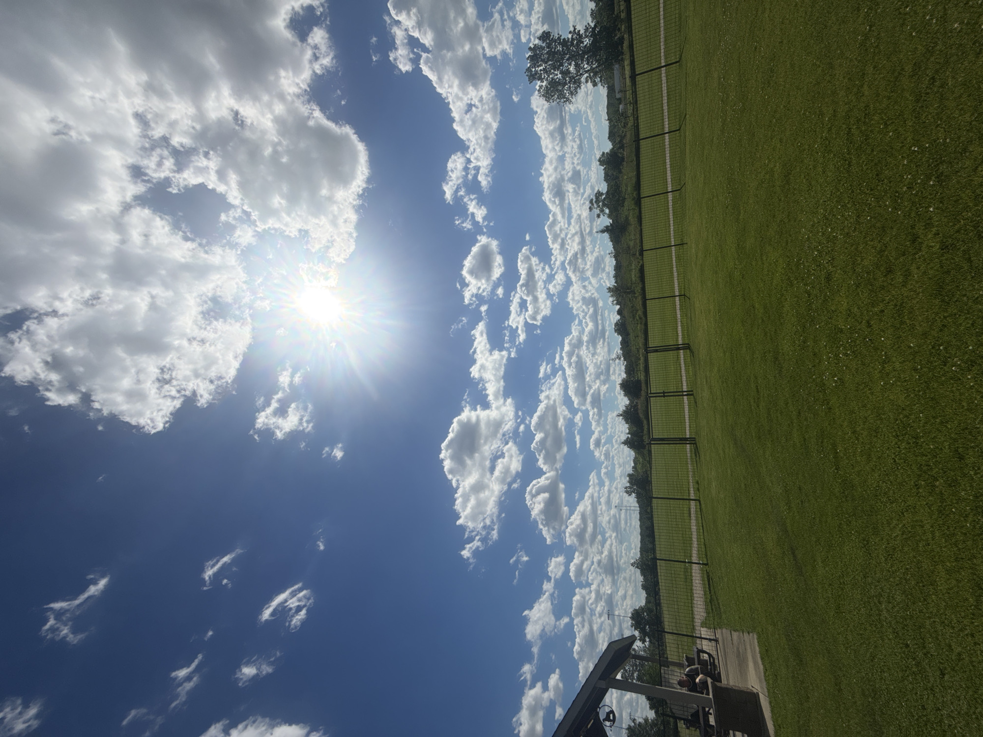 The image shows an outdoor scene with a bright sun and blue sky filled with white clouds. In the foreground, there is a well-maintained green lawn. To the left, a covered pavilion is visible, with a person sitting on a bench underneath. A black fence runs horizontally across the middle ground, separating the lawn from a wooded area and some distant structures. The overall environment appears to be a park or recreational area in Glenview, United States of America.