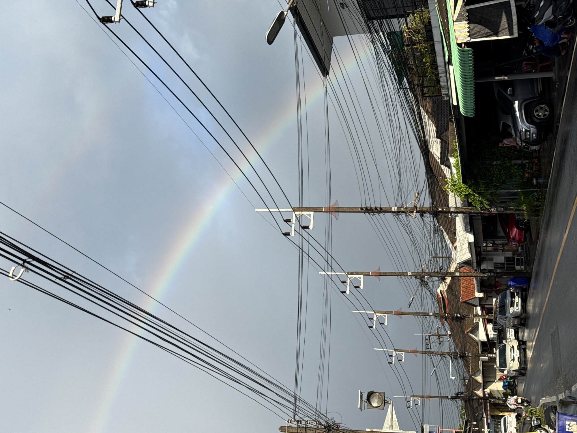 A double rainbow arcs across a gray, overcast sky over a street in Wichit, Thailand, as seen from a low-angle perspective. Power lines crisscross the sky, supported by numerous wooden utility poles, some equipped with streetlights and electrical insulators. The street is wet, suggesting recent rain, and cars are driving along it. The buildings lining the street are a mix of residential and commercial structures with tiled roofs and awnings. On the right side of the street, a parked silver SUV is under an awning. A person wearing a blue garment is seated near parked motorcycles on the right. On the left side of the street, a few cars are parked and moving, and a person is riding a motorcycle. The overall atmosphere is that of a city street under a stormy sky, with the beauty of a double rainbow offering a striking contrast to the urban environment.
