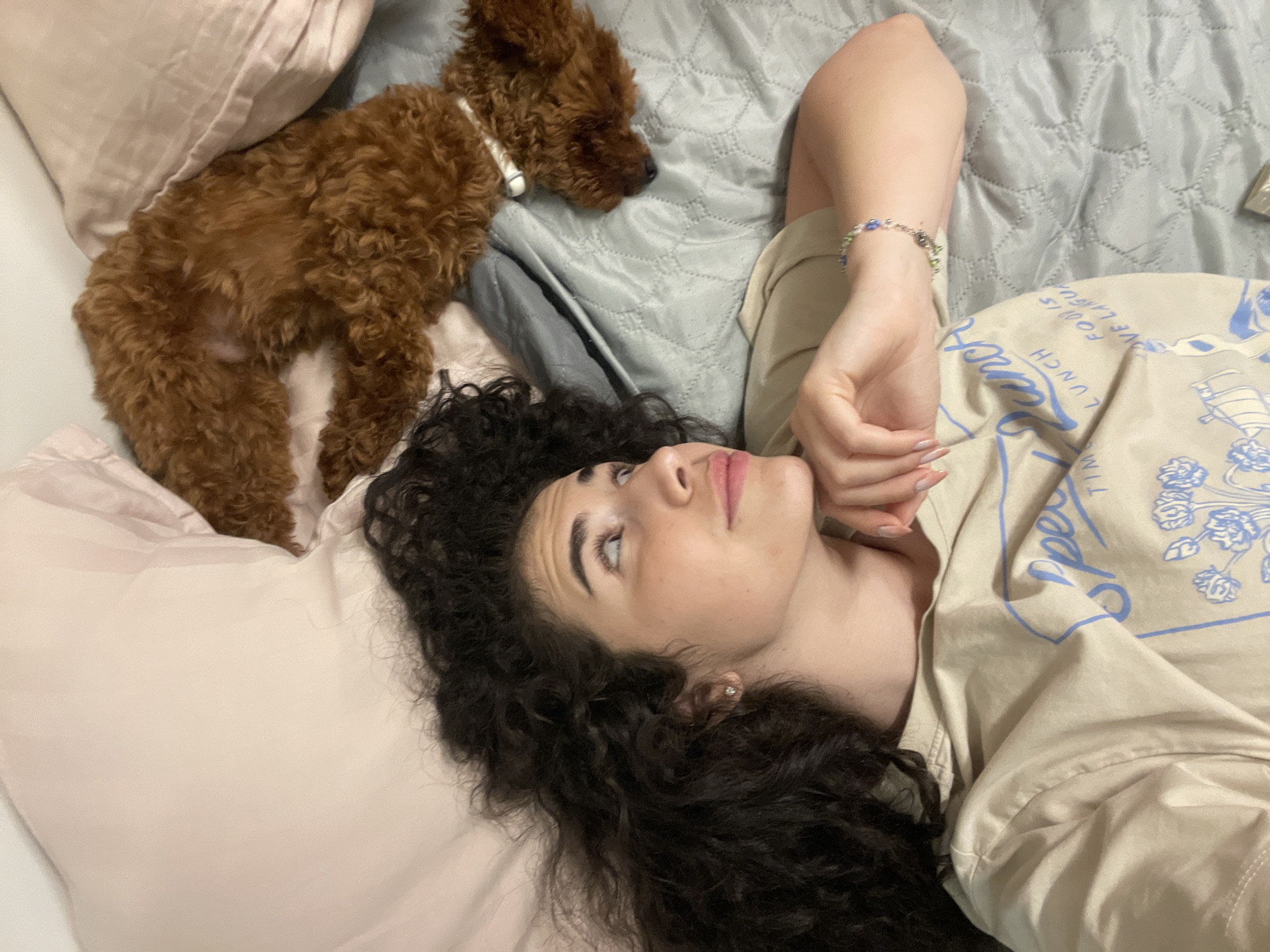 A young woman with dark, curly hair lies on a bed with a small, brown, curly-haired dog. The woman is looking upwards and to the right, her hand resting on her chin. She is wearing a tan t-shirt with a graphic print. The dog is curled up next to her, partially covered by pillows. The bed is made with a gray quilted comforter and light pink pillows. The scene appears to be indoors, likely a bedroom. No specific location in Sofia, Bulgaria is discernible from the image.
