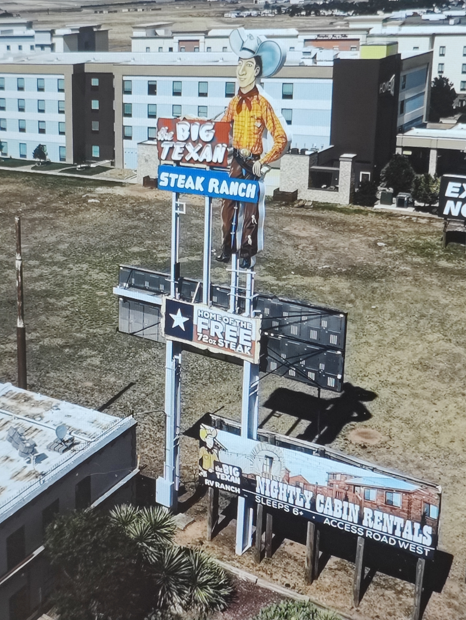 The image shows a roadside scene with large advertisements and buildings in the background. The main focus is a tall sign for "THE BIG TEXAN STEAK RANCH," featuring a cartoon cowboy character. Below this, another sign advertises "HOME OF THE FREE 72oz STEAK." Further down, a sign promotes "NIGHTLY CABIN RENTALS" with the text "SLEEPS 6+" and "ACCESS ROAD WEST." In the background, there are several modern buildings, possibly hotels or apartment complexes, and a large parking lot area. The ground is covered in dry grass and dirt. The overall setting appears to be a commercial area along a highway. The location is not Yarmolyntsi, Ukraine; it is the Big Texan Steak Ranch in Amarillo, Texas, USA.