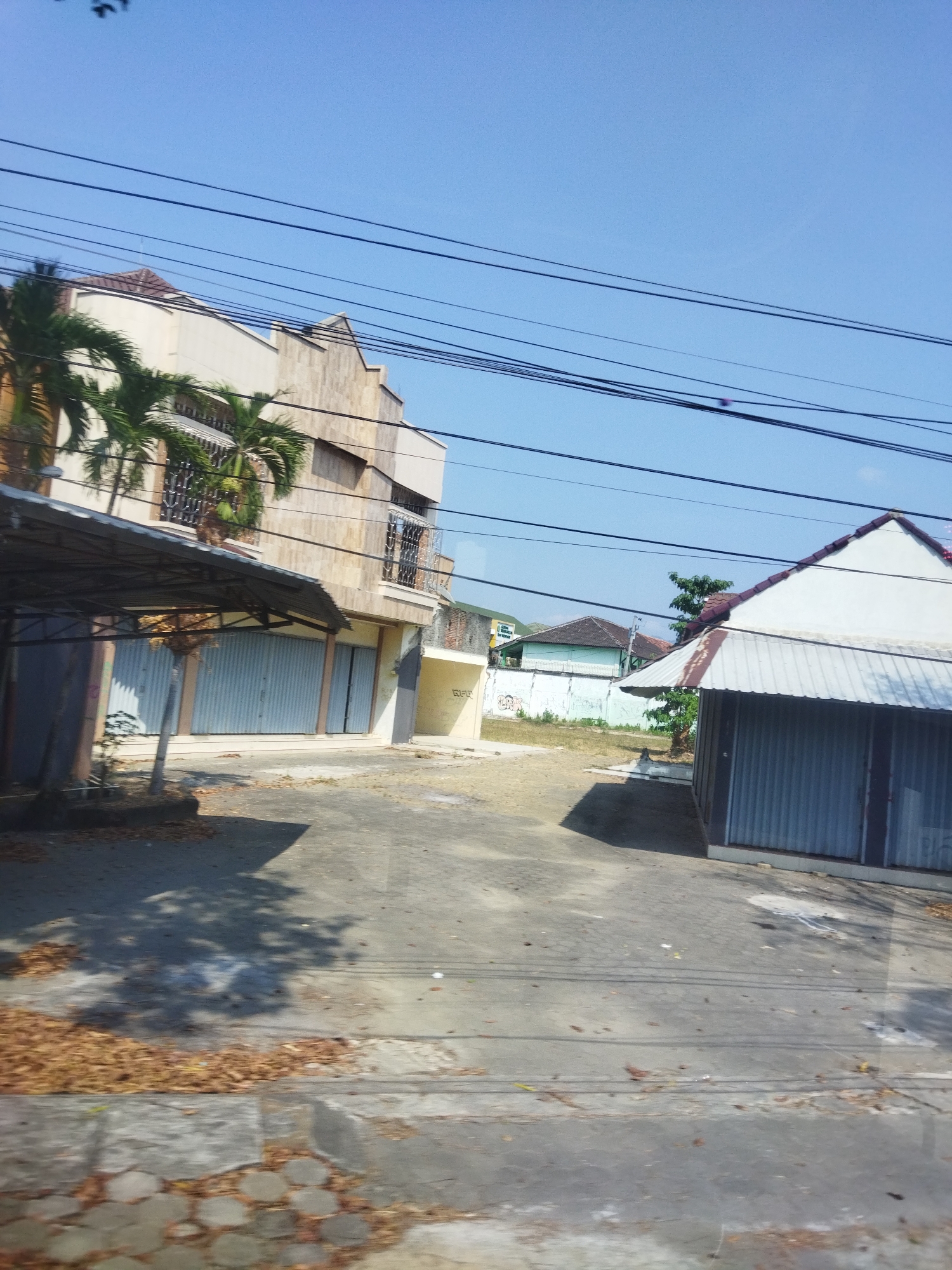 The image captures a street scene in Wonokarto, Indonesia, characterized by a clear blue sky and the presence of several buildings. On the left, a multi-story building with a cream-colored facade and brown tiled roof is visible, featuring windows with decorative metal grilles and balconies. Palm trees are planted near this building. In front of it, a covered area with a metal roof extends outwards, supported by pillars. The lower level of the building appears to house shops with closed metal roll-up doors.
To the right, another building with a white facade and a brown and white corrugated metal roof is situated. This building also has a corrugated metal enclosure in front. In the background, several other buildings with tiled roofs are visible, some with light green and white walls. A tall green tree stands between the two main structures. The ground is paved with asphalt and concrete, with scattered dry leaves and debris. Overhead, multiple power lines crisscross the sky. The overall impression is of a commercial or mixed-use street under bright daylight conditions.