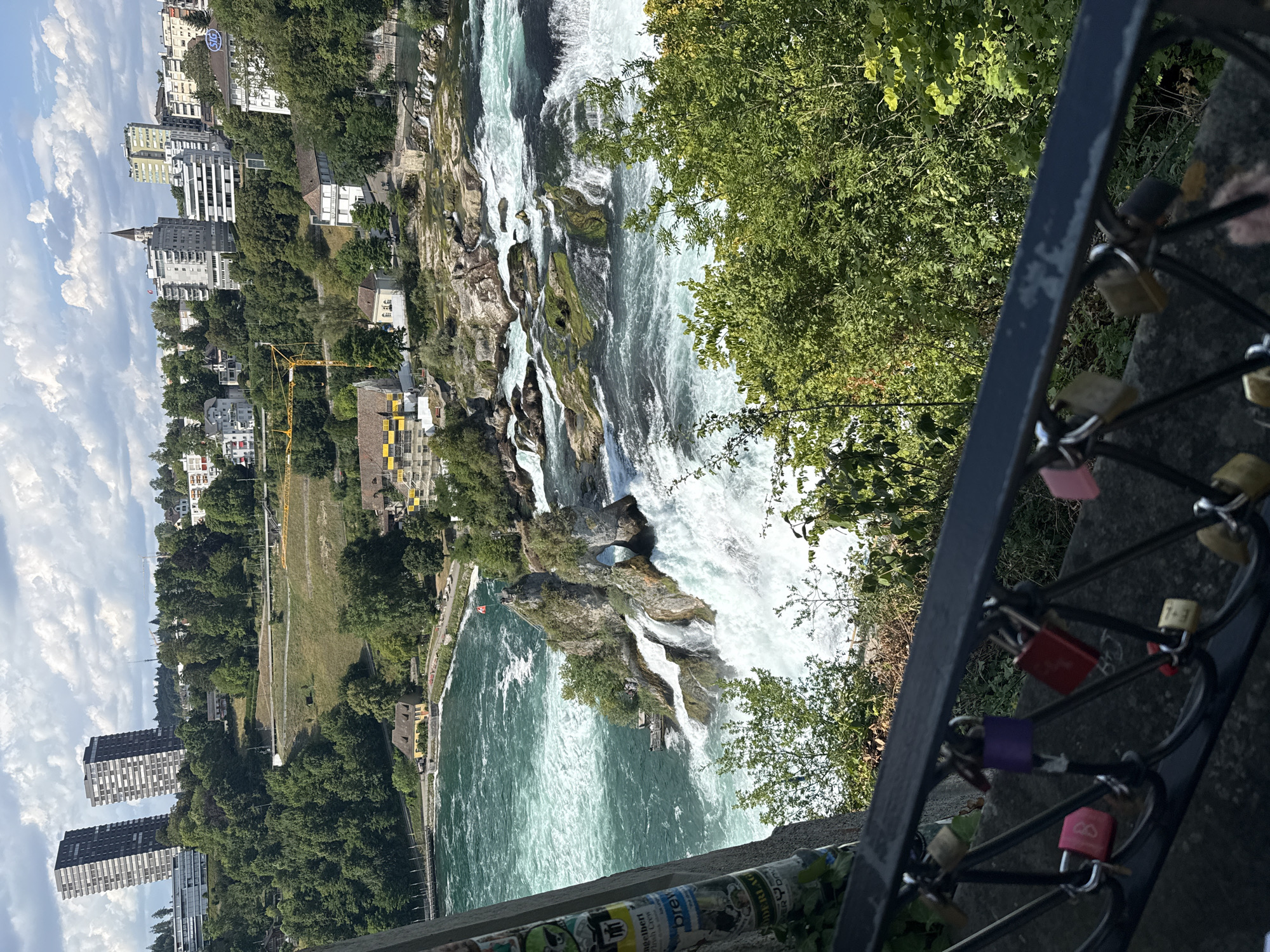 This media file captures a scenic view of the Rhine Falls in Nohl, Switzerland. The image is oriented vertically, looking down towards the powerful waterfall. The main subject is the majestic waterfall itself, with its turbulent white water cascading over large, moss-covered rocks. The water is a vibrant turquoise green where it pools at the base of the falls.
Surrounding the falls, lush green trees and vegetation line the banks. In the background, a city skyline is visible with several buildings, including apartment blocks and older structures. A yellow construction crane is also present, suggesting ongoing development or maintenance in the area. The sky is a bright blue with scattered white clouds, indicating pleasant weather.
In the foreground, a dark metal railing is visible, adorned with numerous colorful love locks of various sizes and hues, including pink, red, purple, and yellow. These locks, attached to the railing, are a common symbol of love and commitment. Some of the locks have numbers or initials inscribed on them.
The overall scene evokes a sense of natural beauty and grandeur, combined with the human touch of the love locks. The lighting suggests it is daytime, likely midday or afternoon. The image conveys a feeling of awe at the power of nature and the romantic sentiment associated with the love locks. Some sticker-like elements with illegible text are visible on a pole or structure near the bottom of the frame.