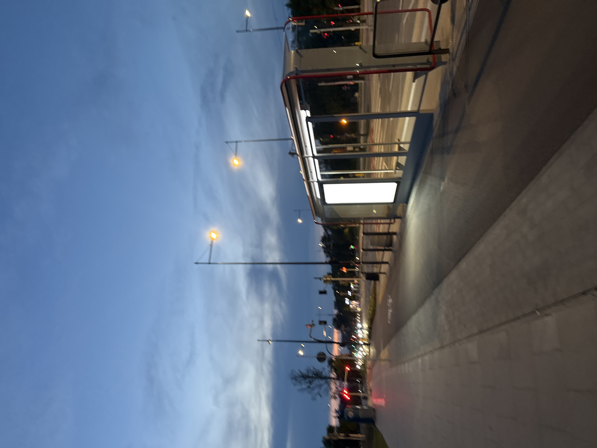 The image depicts a street scene at dusk or early night in an urban environment. On the right side of the frame is a modern bus shelter with a glass enclosure and a white illuminated panel. The bus shelter has red trim and appears to be empty. A paved walkway runs alongside the bus shelter, and beyond that, a road with active traffic is visible, indicated by car headlights and taillights, as well as traffic signals. Streetlights are illuminated, casting a warm glow on the surroundings. The sky is a deep blue with some wispy clouds, suggesting the transition from day to night. The overall impression is one of a quiet evening in a city, with transportation infrastructure prominent.