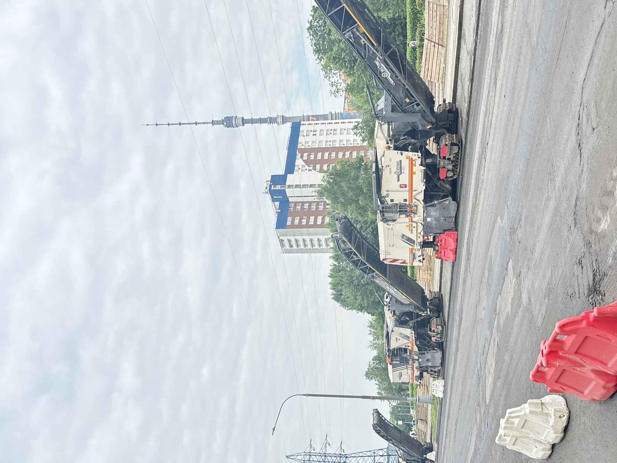 The image depicts a construction site on a cloudy day in Moscow, Russia, likely on an overcast summer day, with a view of the Ostankino Tower in the background. In the foreground, a partially paved road shows signs of recent work, with visible patches of asphalt and some minor damage. Several pieces of heavy construction equipment, specifically milling machines, are positioned on the road, indicating road resurfacing or repair.
Two white and red plastic barriers are placed on the road in the foreground, serving as temporary traffic control. Further down the road, two milling machines are visible. The milling machines are large, industrial vehicles with conveyor belts and rotating drums designed to remove existing pavement. The one in the foreground has "WIRTGEN" branding. Red plastic barriers are also placed near the milling machines.
In the middle ground, behind the construction equipment, there is a row of lush green trees. Beyond the trees, a modern apartment building with white and blue facades stands tall. A tall, slender lattice tower, the Ostankino Tower, is visible in the distance, piercing the overcast sky. A power transmission tower is also present on the left side of the image, adding to the urban industrial landscape. The overall scene suggests ongoing road construction and infrastructure maintenance within a populated urban area of Moscow.
