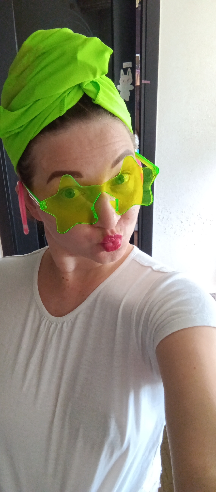 A woman is posing for a selfie. She is wearing a bright green towel wrapped around her head, star-shaped sunglasses with yellow lenses and green frames, and a white t-shirt. She has red lipstick on and is puckering her lips. The background includes a dark door with a cartoon bunny sticker on it and a partially visible white wall. The location is Unknown, Unknown.