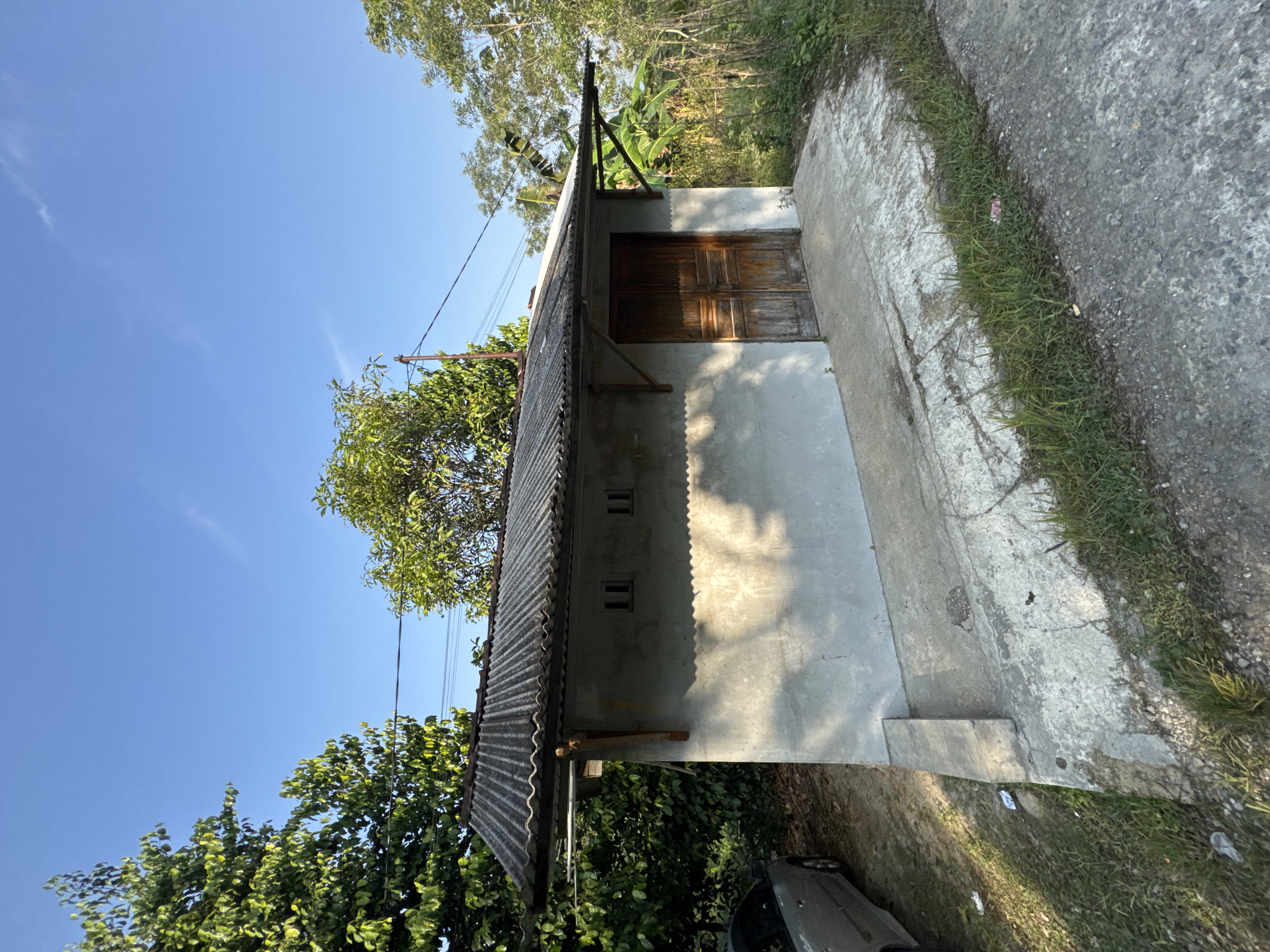 A one-story, weathered concrete building with a corrugated metal roof stands under a clear blue sky in a rural area, possibly Alian, Indonesia. The building has a single set of wooden double doors on its front facade, flanked by two small, square windows. A concrete path leads up to the doors. To the left of the building, a silver car is parked partially visible. Lush green trees and vegetation surround the structure, and power lines stretch across the sky above. The scene appears to be bathed in the warm light of late afternoon or early morning.