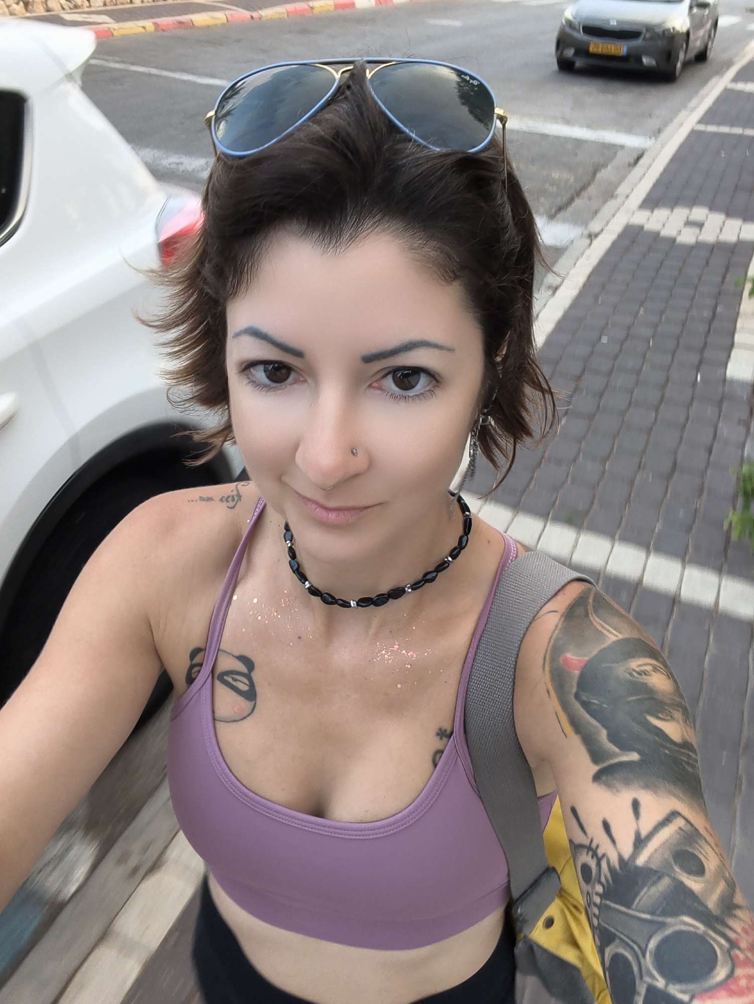 A selfie of a woman with short, dark, wavy hair and brown eyes. She is wearing aviator sunglasses with blue frames, a light purple sports bra, and a black beaded necklace. She has multiple tattoos visible on her left arm and shoulder, including a panda tattoo on her chest and a large, intricate tattoo on her left arm. She is wearing a silver earring in her left ear. The background shows a paved sidewalk with gray cobblestones, a white car on the left, and a dark gray sedan in the background on the street, suggesting an urban setting. The scene is consistent with a location in the city of Nahariyya, Israel.