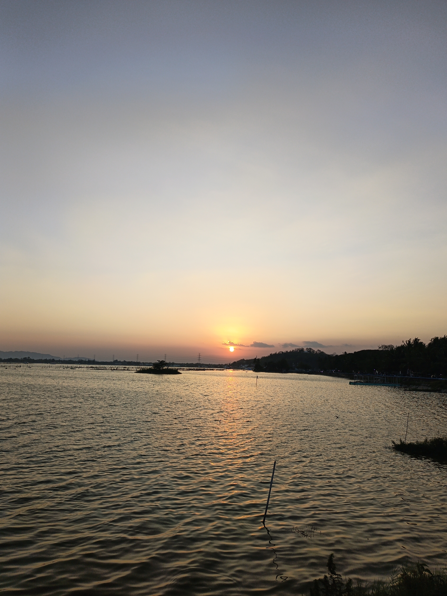 A serene sunset scene unfolds over a tranquil body of water, likely a lake or reservoir, in Krakitan, Indonesia. The sky transitions from a pale blue at the top to soft hues of orange and yellow as it approaches the horizon, where the sun, partially obscured by a thin cloud, casts a warm, golden glow. The water's surface is rippled, reflecting the warm colors of the sky. In the distance, silhouetted against the sunset, are power lines and what appears to be a low-lying island or marshy area dotted with vegetation and poles. To the right, the shoreline is lined with trees and what seem to be small structures or boats docked. A few vertical poles are visible emerging from the water, some in the foreground and others further out. There are no visible people or specific actions depicted. The overall atmosphere is peaceful and natural.