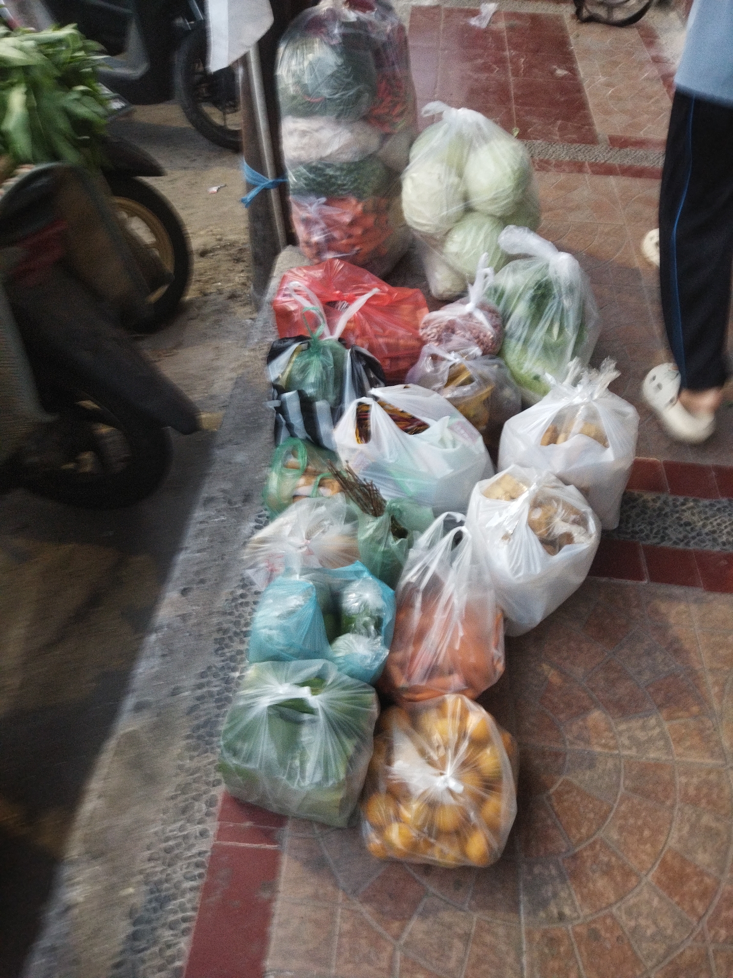In Banjar, Indonesia, an outdoor marketplace scene unfolds. A motorcycle, partially visible on the left, rests on a stone-paved walkway. Adjacent to it, a vendor displays an assortment of produce, packaged in clear plastic bags. Vegetables like cabbage, leafy greens, and root vegetables are neatly arranged. Scattered among the vegetables are bags of what appear to be nuts or small fruits. The ground is a patterned tile. A person's legs, clad in dark pants and white slip-on shoes, are visible on the right side of the frame, indicating their presence near the market stall. The lighting suggests it is daytime.