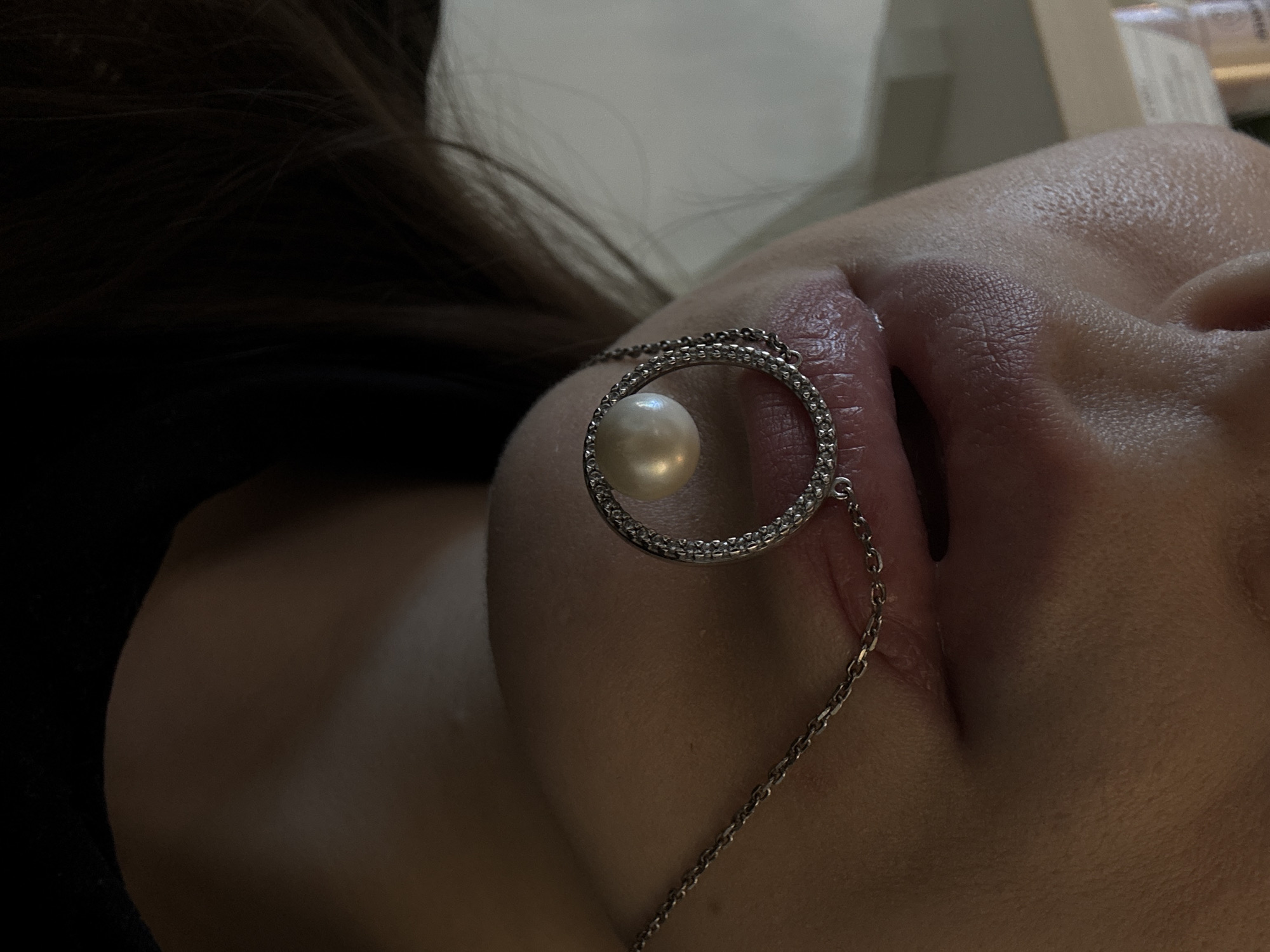 The image is a close-up, medium shot of a person's mouth and chin. The person has on a necklace that hangs from their mouth and has a circular pendant with a pearl in the center. The necklace appears to be made of silver with small stones encrusting the circular pendant. The person's lips are slightly parted, revealing the necklace. The background is blurred and out of focus. There is no indication of location in the city of Unknown, Russia.