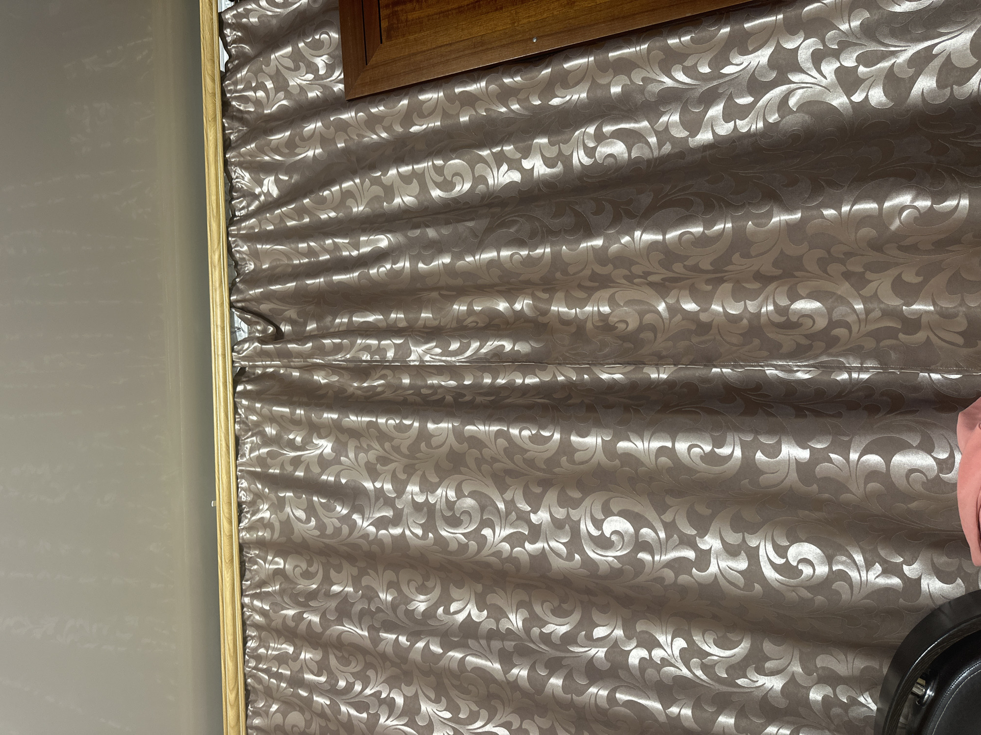 The image shows a close-up view of a patterned curtain. The curtain is a neutral, shimmering color with a raised floral or paisley design. It is hanging from a wooden rod at the top of the frame. To the right of the curtain, a portion of a wooden furniture piece, possibly a cabinet or door, is visible. The ceiling above the curtain appears to be a textured white material. A dark-colored object, possibly the back of a chair, is visible in the bottom left corner of the frame. A piece of pink fabric is also visible in the bottom center. The scene appears to be indoors, in a room with decorative elements. Without further context, it is impossible to determine the specific location within Загородный, Russia.