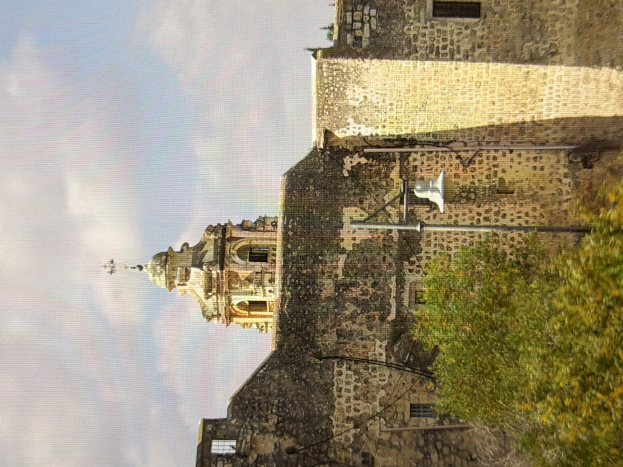 The image shows a weathered stone building with a bell tower topped by a cross. The tower has arched windows and ornate architectural details. The building's walls are constructed of rough-hewn stones, some of which appear to be crumbling. A modern street lamp with a white bell-shaped fixture stands in front of the building. Green foliage is visible in the foreground, and the sky above is partly cloudy. There are no people or animals visible in the image. The building's architecture and material suggest it is an old religious structure, possibly a church or monastery, located in a historical district of Kyiv, Ukraine.