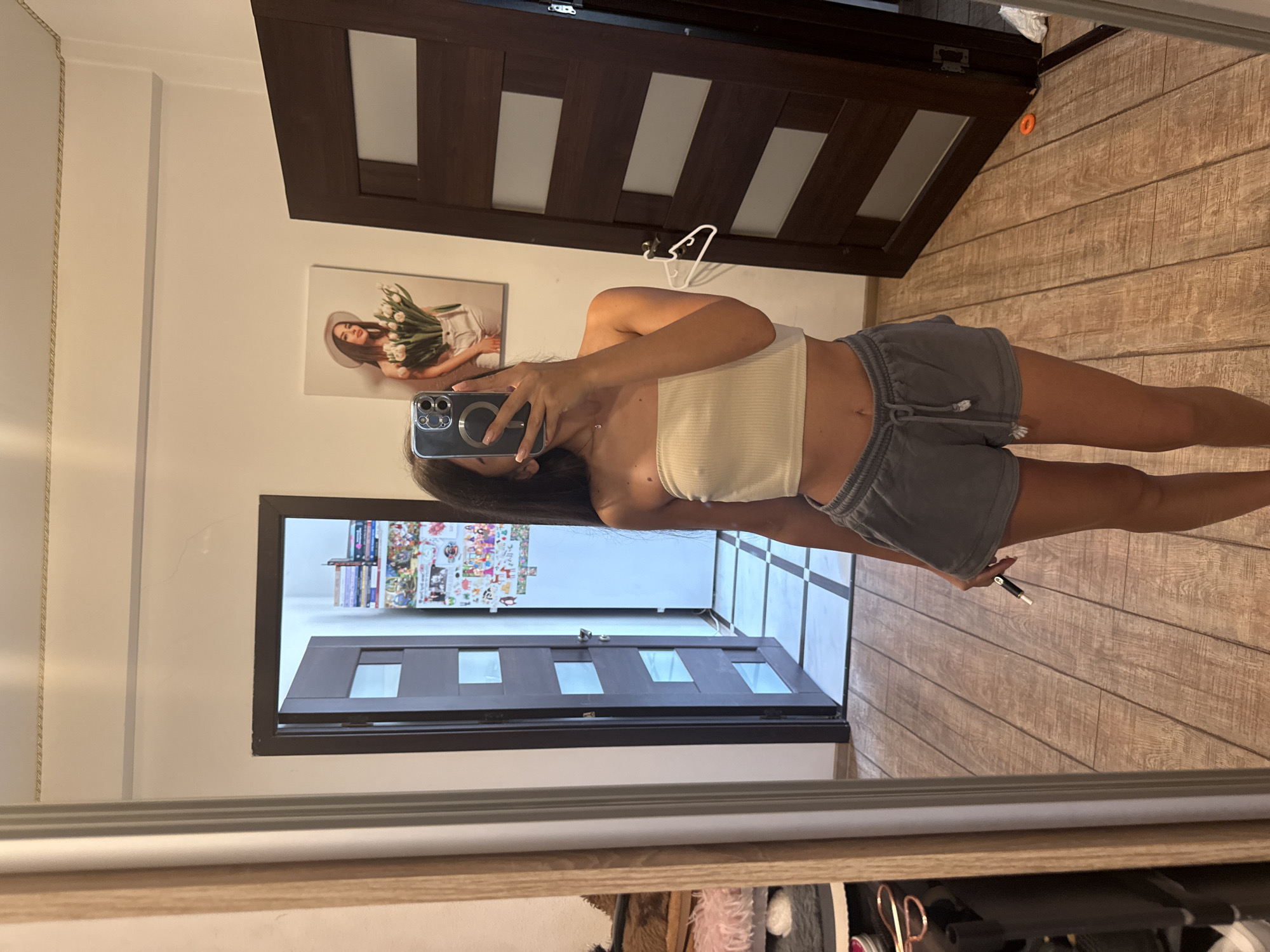 A young woman, with long dark hair, is taking a mirror selfie in what appears to be a bedroom or hallway. She is wearing a white strapless crop top and gray shorts. She is holding a smartphone in her right hand to take the photo. On the wall behind her, there is a framed picture of a woman holding flowers, and to her left, a door with glass panels. The floor is tiled with a wood-like pattern. To the left of the frame, a mirror reflects part of the room and a collection of shoes and other items. The overall scene suggests a casual, domestic setting. The specific location in Chișinău, Moldova, cannot be determined from the visual information provided.