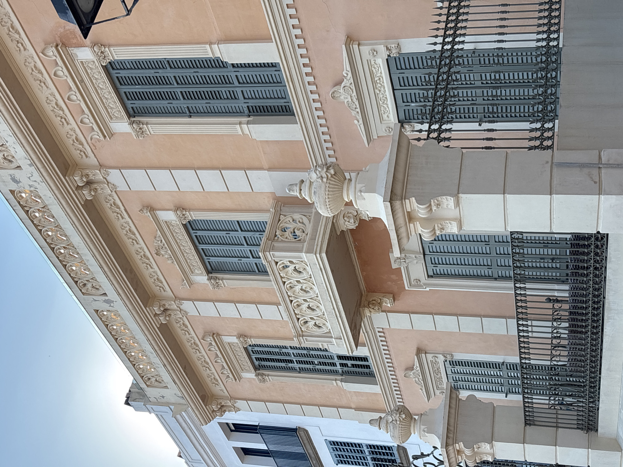This is a vertical, eye-level, outdoor photograph of the upper facade of a building, likely a historic apartment building or hotel, in Calella De Palafrugell, Spain. The building's exterior is painted a light peach color with white trim and intricate decorative molding in a light beige color. The architectural details include ornate carvings above and around the windows, fluted pilasters, and decorative urns or finials. Several windows are visible, each fitted with dark gray louvered shutters. Some windows are protected by decorative black wrought-iron balconies. The sun is bright, casting a lens flare in the upper left corner and suggesting it is daytime, likely late morning or early afternoon, with clear, bright weather. The image is angled upwards, giving a sense of looking towards the sky, which is a pale blue. There is no visible text in the image. The scene conveys a sense of European coastal charm and architectural beauty.