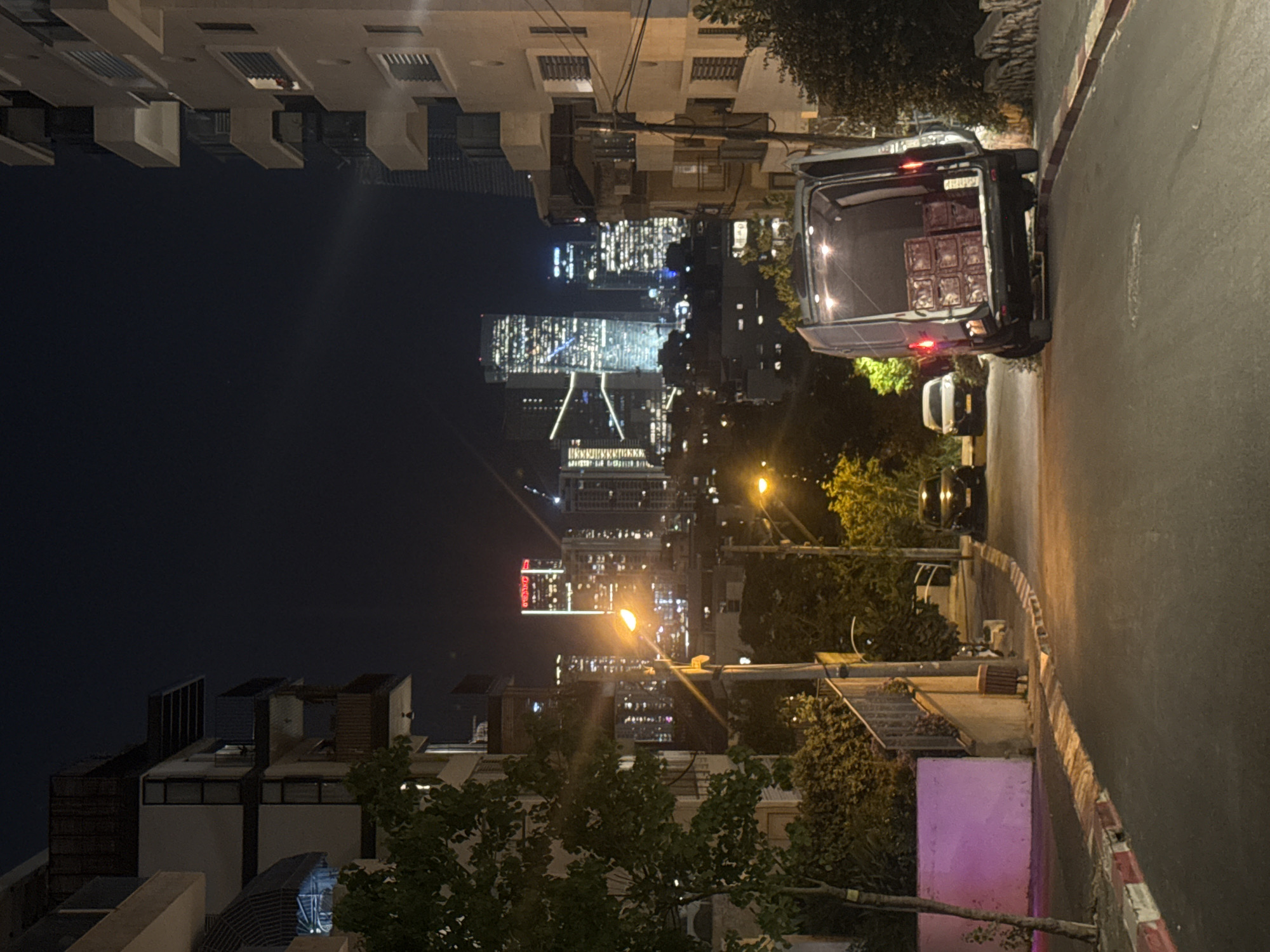 The image shows a nighttime view of Givatayim, Israel, from street level. Tall buildings with illuminated windows dominate the skyline. A street curves to the right with a parked van, its back open revealing stacked crates. A row of buildings lines the left side of the frame, with trees obscuring some of the lower levels. Streetlights cast a glow on the scene.