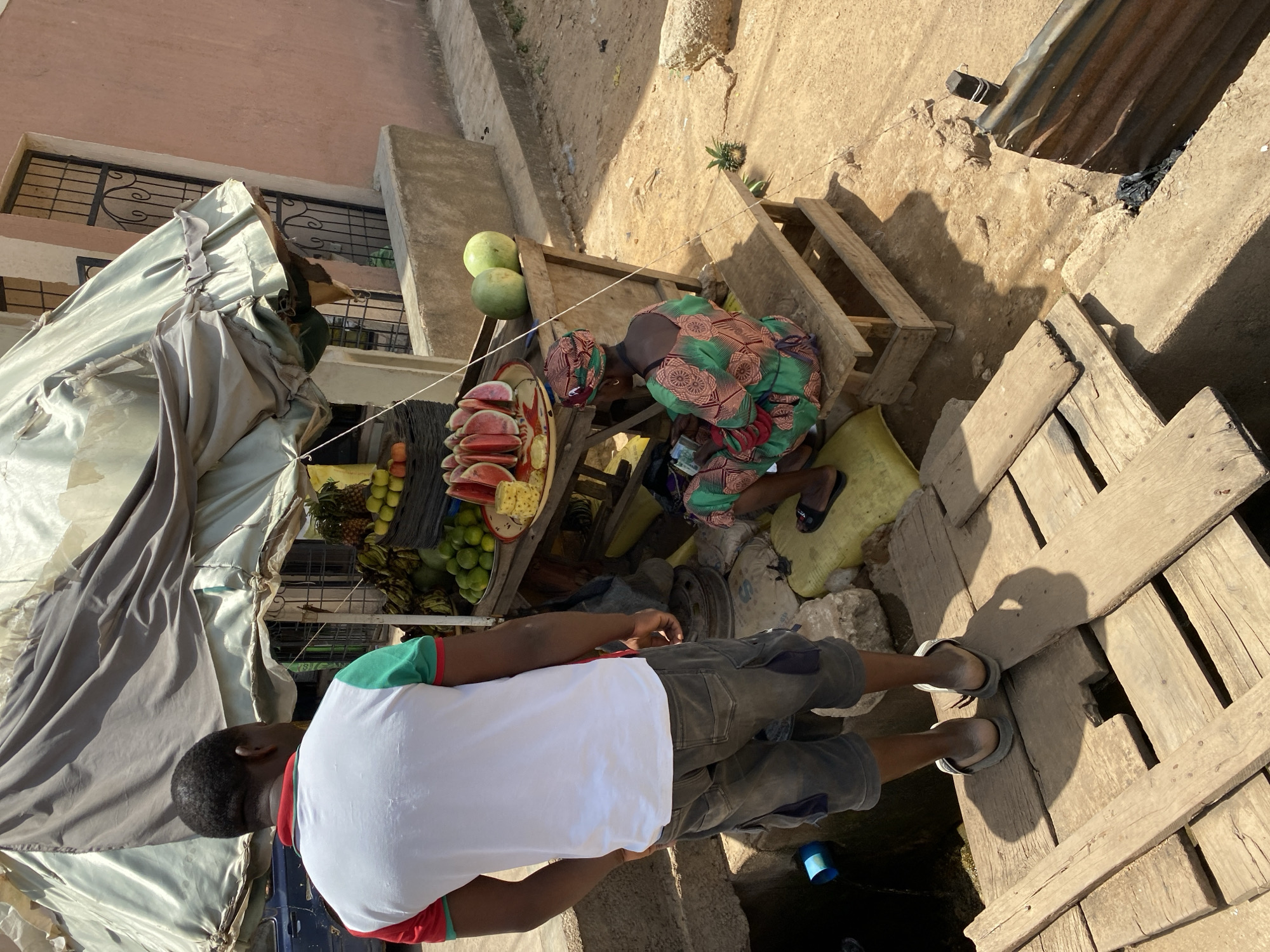 In Bukuru, Nigeria, a man in a white t-shirt, gray shorts, and sandals stands on a wooden platform next to a street-side fruit stand. The stand, covered by a gray tarp, displays watermelons, pineapples, limes, and other fruits. A woman in a patterned green dress and head covering sits behind the stand, appearing to be handling money. The location is outdoors, with a building visible in the background and varying ground textures of concrete and dirt.
