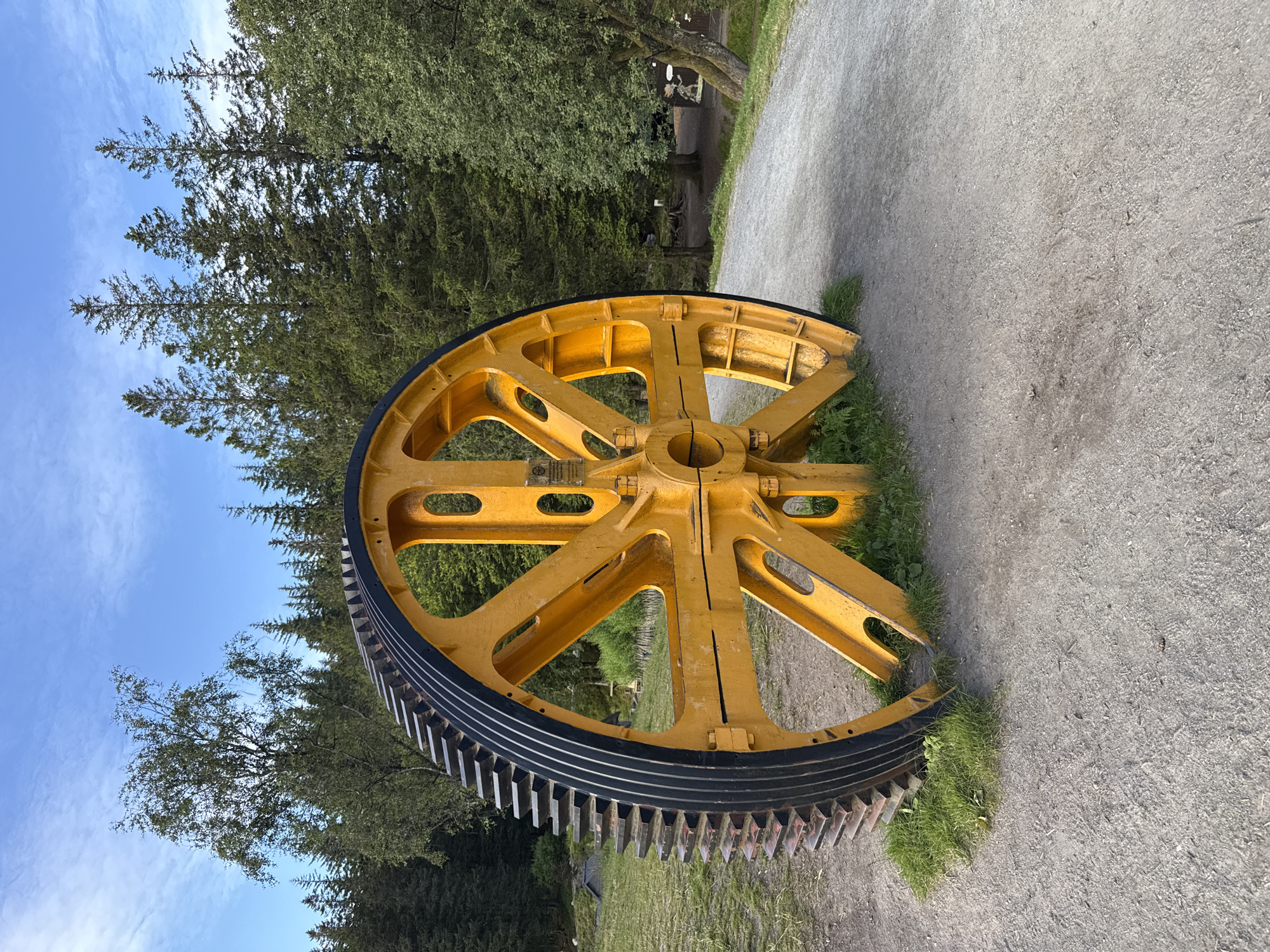 An oversized, bright yellow gear, likely a remnant of industrial machinery, stands prominently in a grassy area next to a gravel path. The gear has a toothed outer edge and a spoked interior with circular cutouts. Behind it, a dense forest of evergreen trees stretches across the landscape under a bright blue sky with scattered white clouds. To the left, a deciduous tree with lighter green foliage is visible. The scene is part of the Fløyen mountain, accessible via the Fløibanen funicular in Bergen, Norway, a popular tourist destination offering panoramic views of the city and surrounding fjords. There are no people or animals present in the image. The lighting suggests it is daytime, with clear visibility.