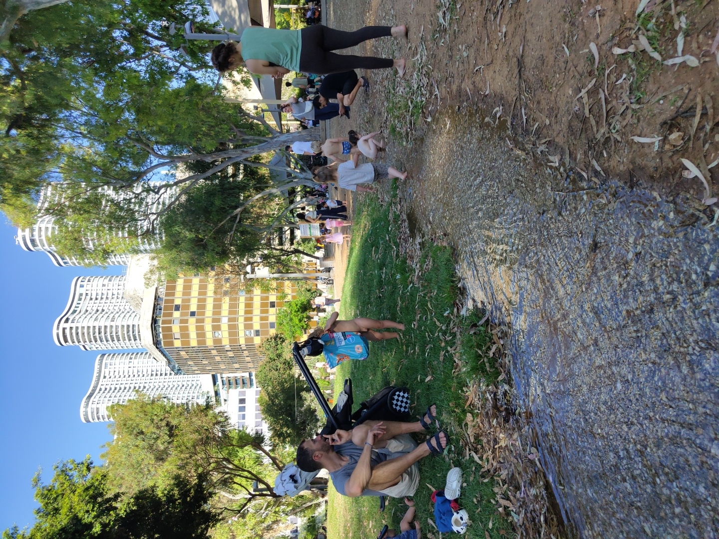 In Tel Aviv, Israel, on a sunny day, a park scene unfolds with a shallow, rocky stream flowing in the foreground. On the grassy bank to the left, a man sits with his legs crossed, wearing a grey t-shirt and beige shorts, with sandals on his feet. Beside him, a stroller is parked. A small child, partially visible and naked, stands near the stroller holding a light blue bag.
Further into the park, several people are scattered. A woman in a green tank top and black leggings stands facing away from the camera, while a man crouches down near her. Children are also present, some playing in or near the stream. One child, barefoot and wearing grey shorts, walks into the stream. Another child, also barefoot, is crouched down in the shallow water. In the background, a building with a yellow facade is visible, and towering white, curved buildings are seen behind it. The trees provide shade, and the ground is covered with dry leaves. The sky is clear and blue.