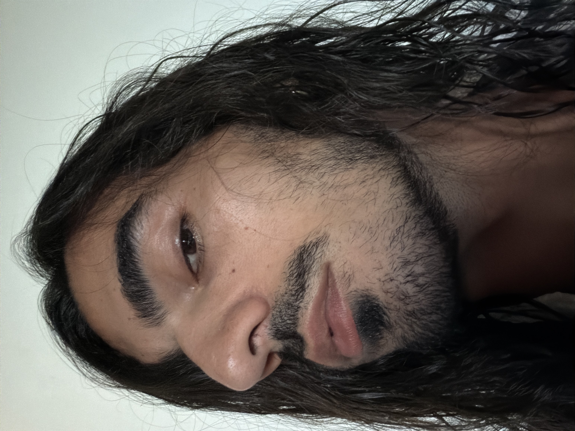 This is a close-up portrait of a person, rotated 90 degrees counter-clockwise, against a plain, light-colored background. The subject, appearing male, has long, dark, wavy hair that looks wet or styled with a sleek, shiny texture, framing their face and flowing out of the frame. Loose, fine strands of hair are visible around the temples and hairline.
The person has thick, dark, and well-defined eyebrows. Their visible right eye is dark brown and appears to have a reflective sheen, possibly from moisture. They have a prominent nose and full lips, with a dark, short beard and mustache covering the upper lip, chin, and jawline. The skin has a warm tone and appears smooth and somewhat dewy or moist, especially around the nose and forehead, reflecting the soft, diffused lighting. The expression is neutral, possibly pensive or relaxed.
The image focuses exclusively on the individual's head and upper face, offering no discernible objects or details about the immediate scene. No specific activity is depicted beyond posing for the portrait. The time of day and weather are indeterminate. No text is visible. While the location context provided is North Kuta, Indonesia, no specific geographical cues are present in the image itself.