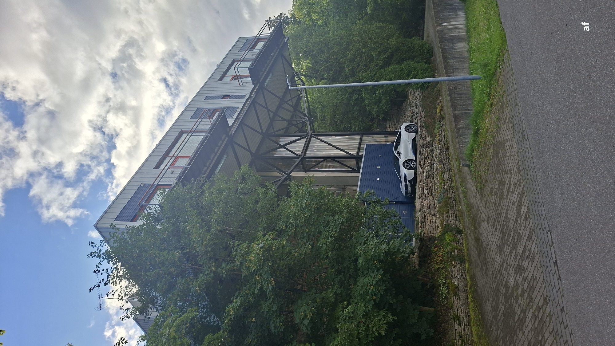 In Tabasalu alevik, Estonia, a white BMW i8 sports car is parked on a stone-lined terrace in front of a modern multi-story building. The building features a metallic facade, balconies with orange railings, and large windows. A distinctive cantilevered structure with a metal truss system extends from the building, supporting an upper level. The area is surrounded by lush green trees and foliage. The ground in the foreground is paved with interlocking bricks, and a concrete retaining wall is visible to the right, bordered by grass. The sky is partly cloudy with patches of blue.