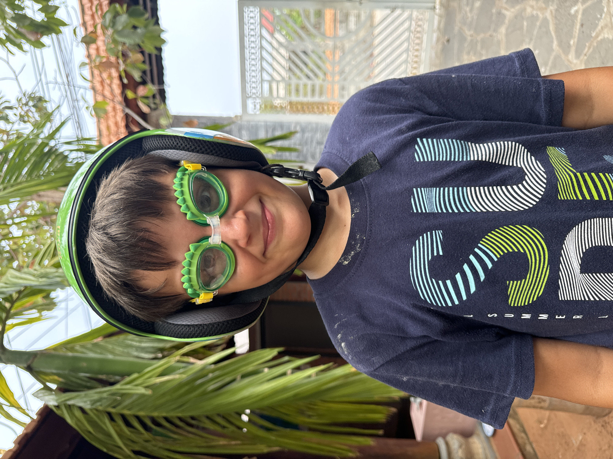 A child is centered in the frame wearing a green helmet and green dinosaur-themed goggles. The child is also wearing a dark blue t-shirt with white and green lettering. The background includes lush greenery, a building with brown roof tiles, and a decorative white metal gate. The location is Mũi Né, Vietnam. The photo is taken at an angle, with the child and foreground foliage appearing on the left side, and the background elements on the right.