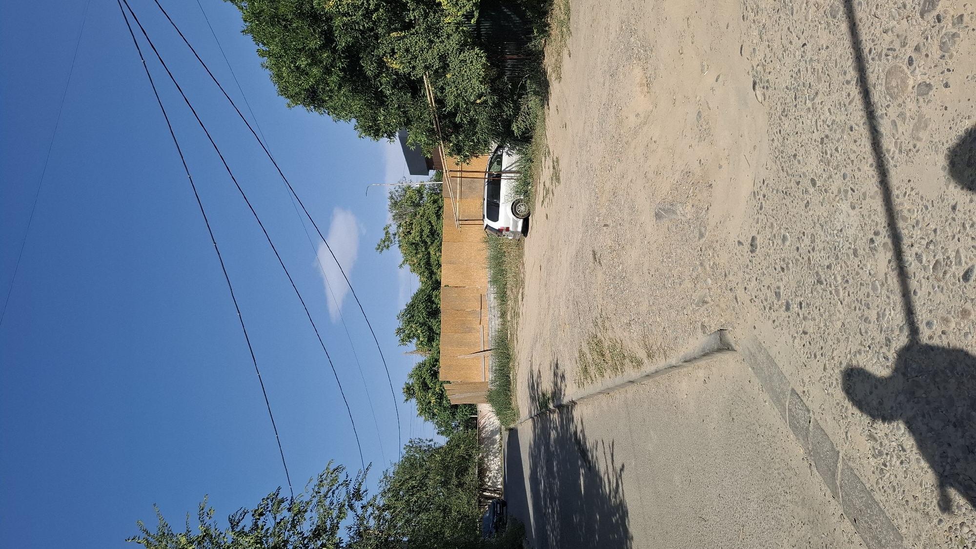 The image depicts an outdoor scene on a sunny day, likely in Shymkent, Kazakhstan, given the context provided. In the foreground, a shadow of a person holding a phone is cast on a paved road with exposed pebbles and a concrete curb. To the right of the road is a dirt and gravel lot.
In the middle ground, a beige wooden fence, possibly under construction or temporary, spans across a significant portion of the scene. This fence is built on a low concrete foundation and is surrounded by overgrown green grass and weeds. A white van is parked on the dirt lot to the right of the fence, partially obscured by a tree and a makeshift lean-to structure with a bamboo frame. Further to the left, a dark-colored car is visible on the paved road, driving away from the viewer.
The background features a dense line of green trees and foliage, suggesting a residential or semi-urban area. Above, a clear blue sky is dotted with a few wispy white clouds. Several dark electrical wires stretch across the sky, contributing to the urban or suburban atmosphere. The overall impression is one of a warm, bright day in a developing or somewhat unkempt area.