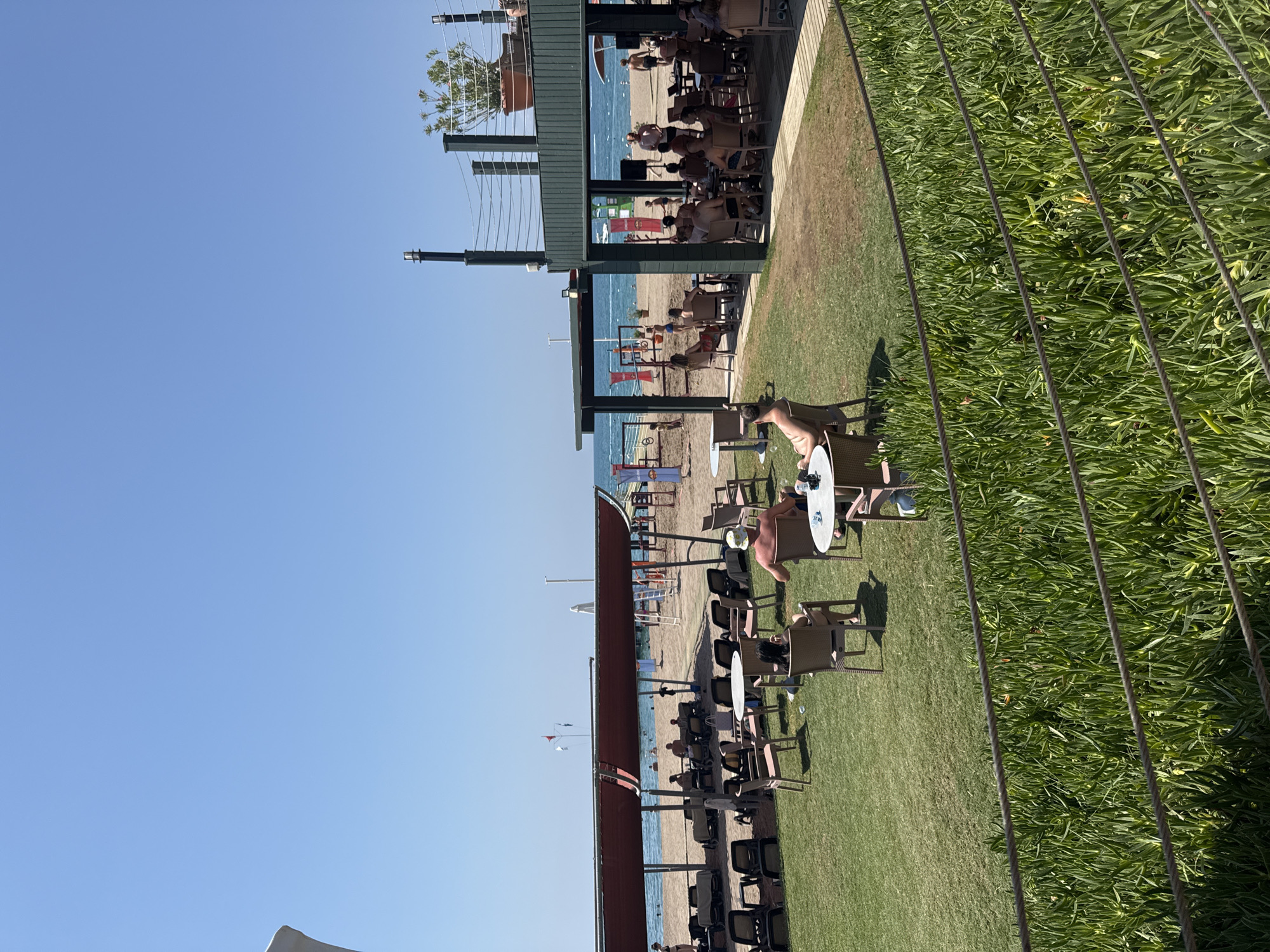 This is a wide shot of a resort beach in Aksu, Turkey, on a clear, sunny day. In the foreground, a lush green lawn is bordered by a rope fence. Several people are seated at small, round tables on the lawn, some in chairs and others on lounge chairs. A covered walkway or outdoor seating area with a dark green roof and pillars is visible to the right, with more tables and chairs, and people dining. Beyond this area is a sandy beach with numerous lounge chairs and umbrellas, and people sunbathing and walking. The sea is visible in the background, with small waves. In the far background, beach activities or games appear to be set up, with colorful flags and structures. The overall atmosphere is relaxed and recreational, typical of a beach resort.