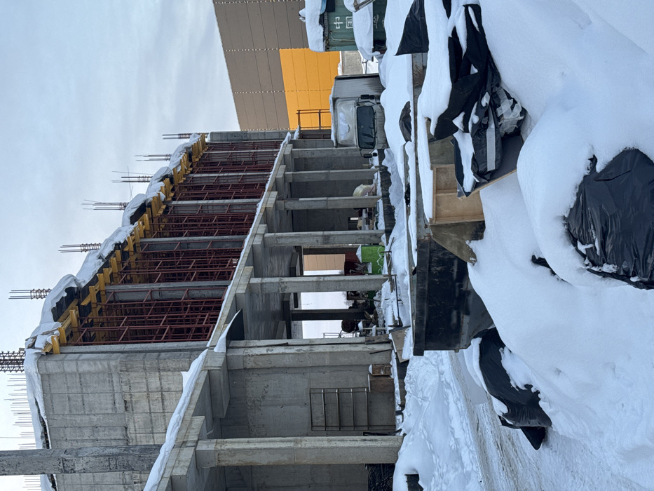 The image displays a winter construction site located in L'vovskiy, Russia, under an overcast, light grey sky. The scene is heavily covered in white snow.
In the foreground, large piles of snow conceal dark debris, some wrapped in black plastic sheeting. A dark-colored, open-top dumpster is partially visible in the center, filled with snow and various materials. To its right, wooden construction elements are partially obscured by snow.
The midground is dominated by an unfinished multi-story concrete building structure. Its exposed concrete framework includes numerous columns supporting multiple floor slabs. The upper visible floor features red metal scaffolding supporting yellow formwork panels along a significant portion of its length, with exposed rebar protruding from the top edge, indicating ongoing construction. Snow accumulates on the concrete slabs and formwork. Beneath the elevated section of the building, a green portable toilet, a white box truck, and a blue shipping container marked with Chinese characters are discernible amidst various construction materials and equipment. A metal shelving unit is visible under the structure on the left.
In the background, a large, rectangular building with a brown paneled facade featuring a prominent yellow section is visible to the right of the concrete structure. No individuals are present in the scene.